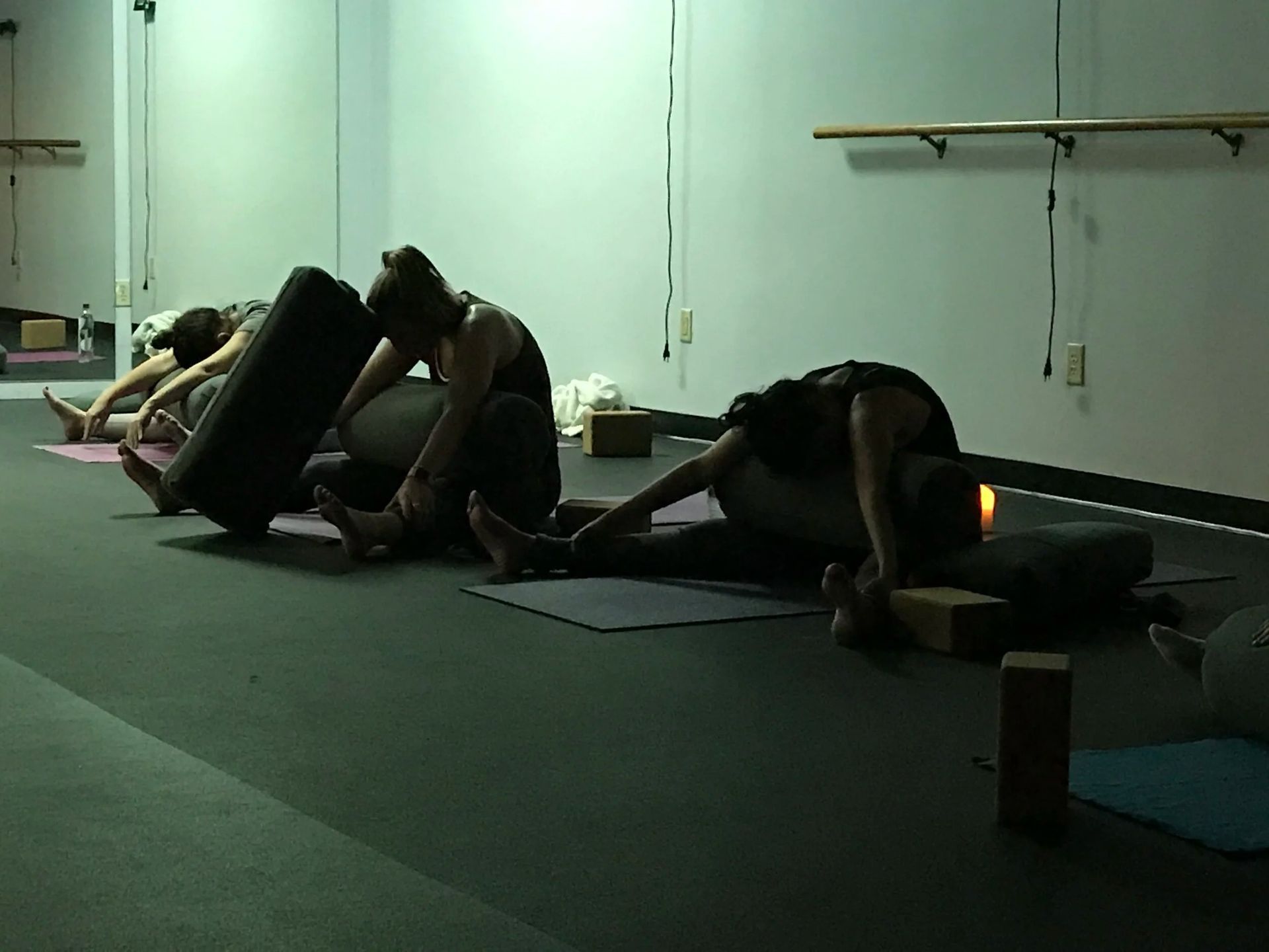 People in a dim yoga studio practicing a seated forward fold pose using cylindrical bolster cushions.