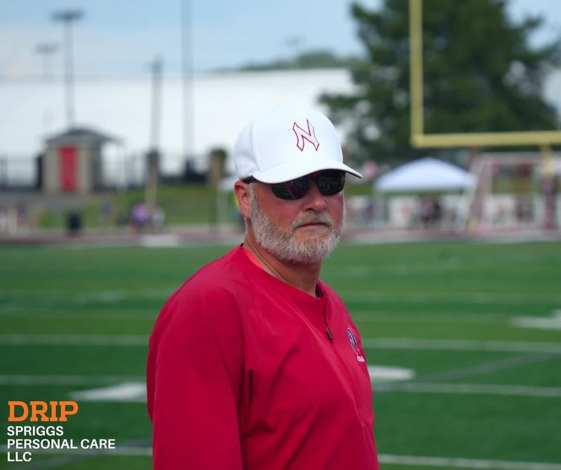 Man in red shirt and white cap standing on a sports field, wearing sunglasses