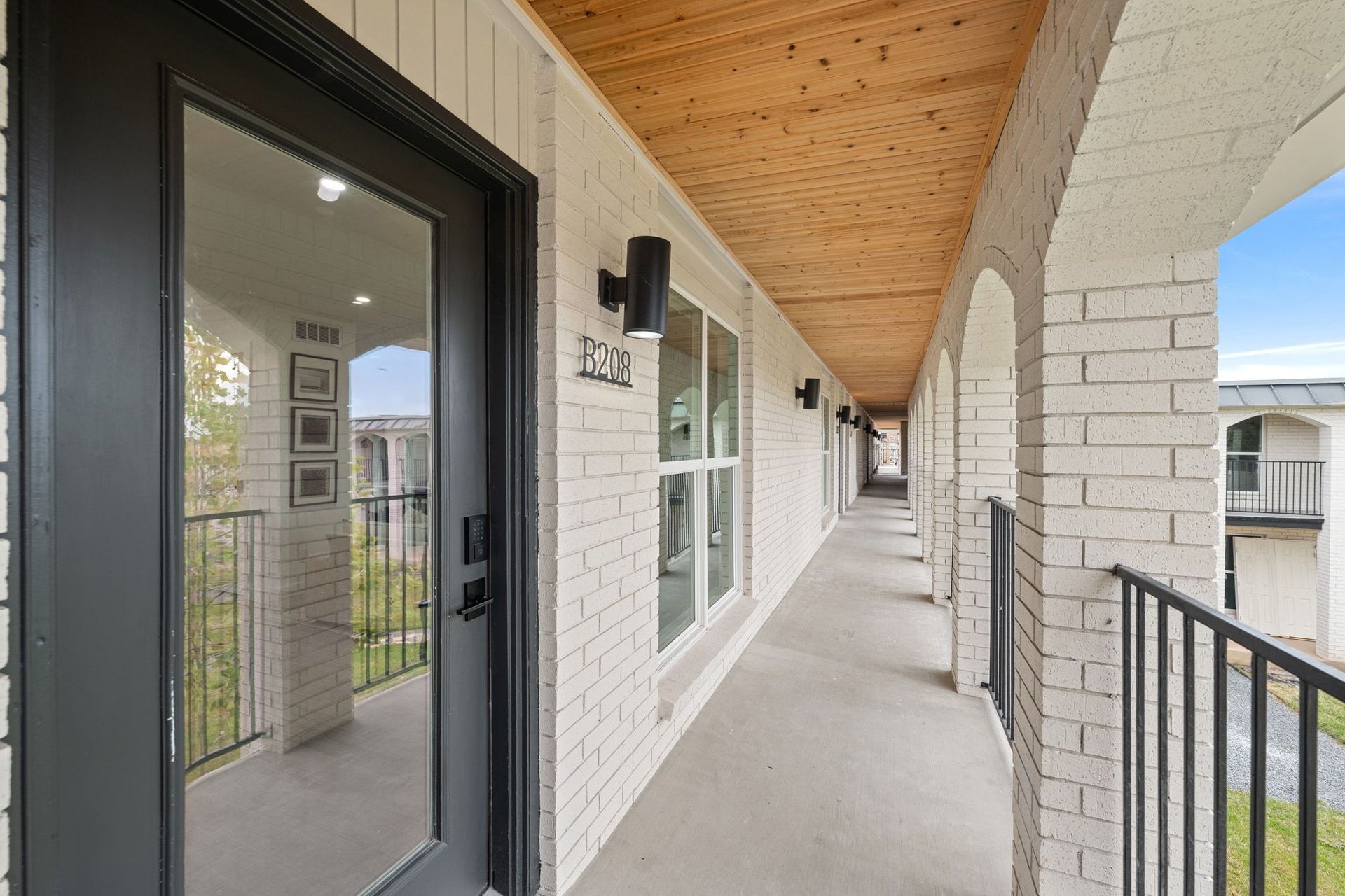 Covered apartment walkway with white brick walls, black railings, and glass doors under a wood ceiling