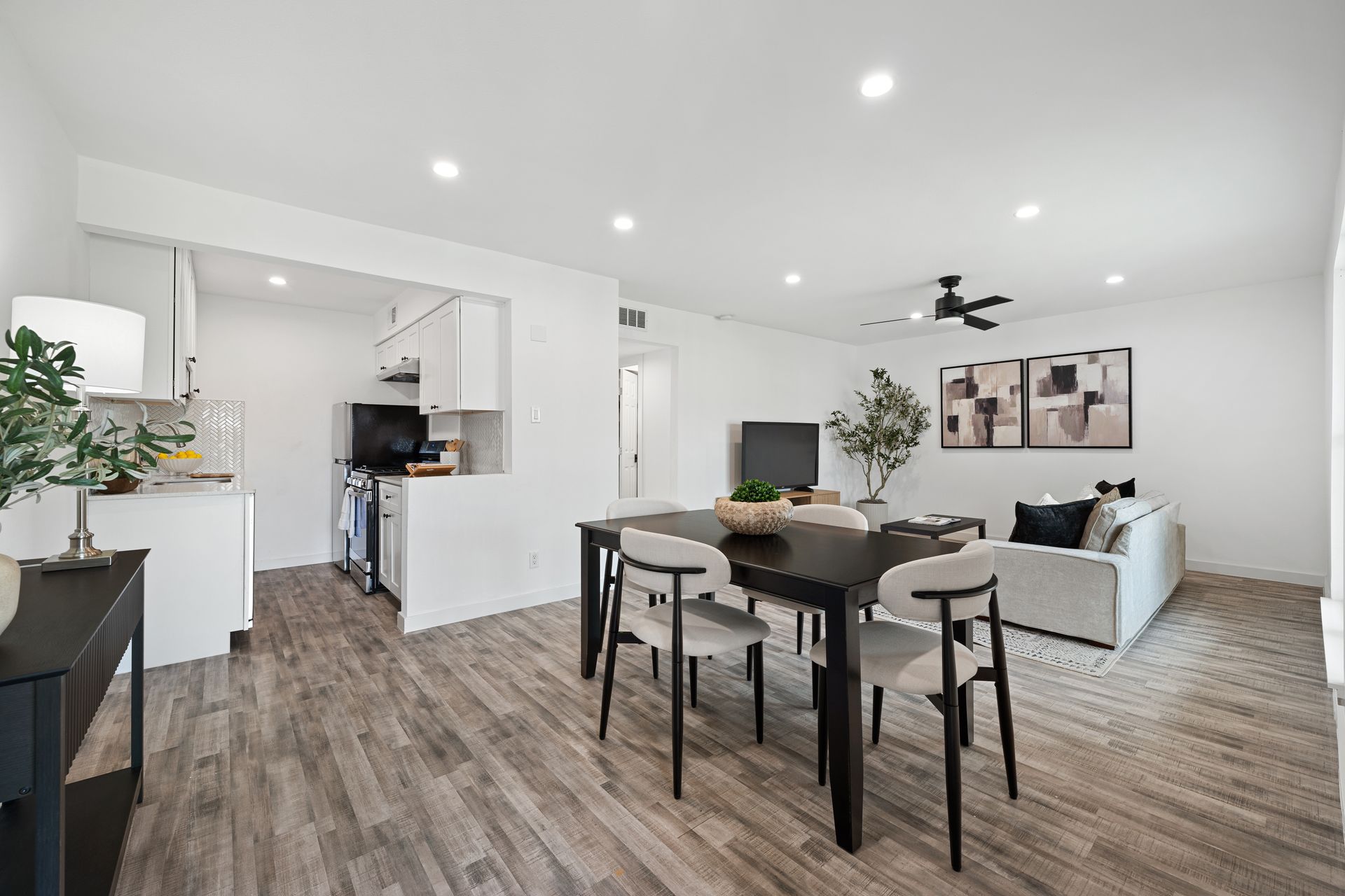 Bright open-concept living room and dining area with kitchen, white walls, wood floors, and black dining table.
