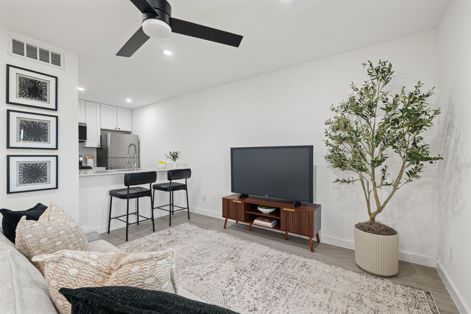 Modern living room with sofa, TV, bar stools, ceiling fan, and a potted tree beside a kitchen area