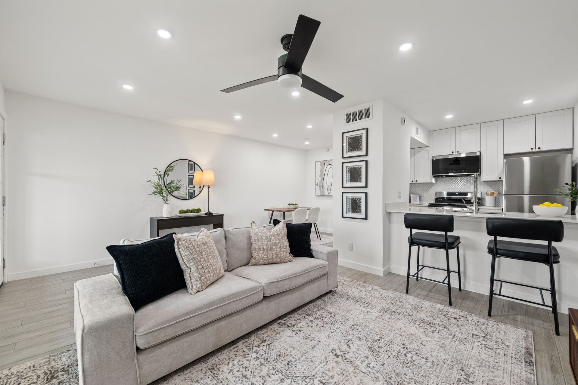 Bright modern living room with gray sofa, black ceiling fan, and open kitchen with bar stools.