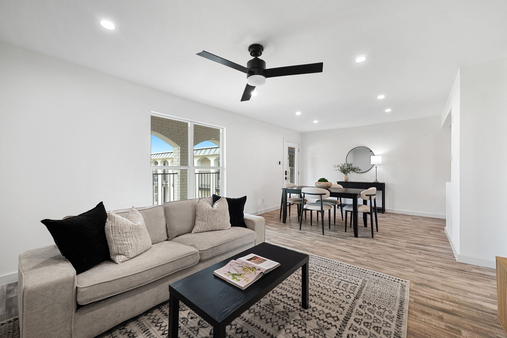 Bright modern living room with beige sofa, black coffee table, dining area, and ceiling fan