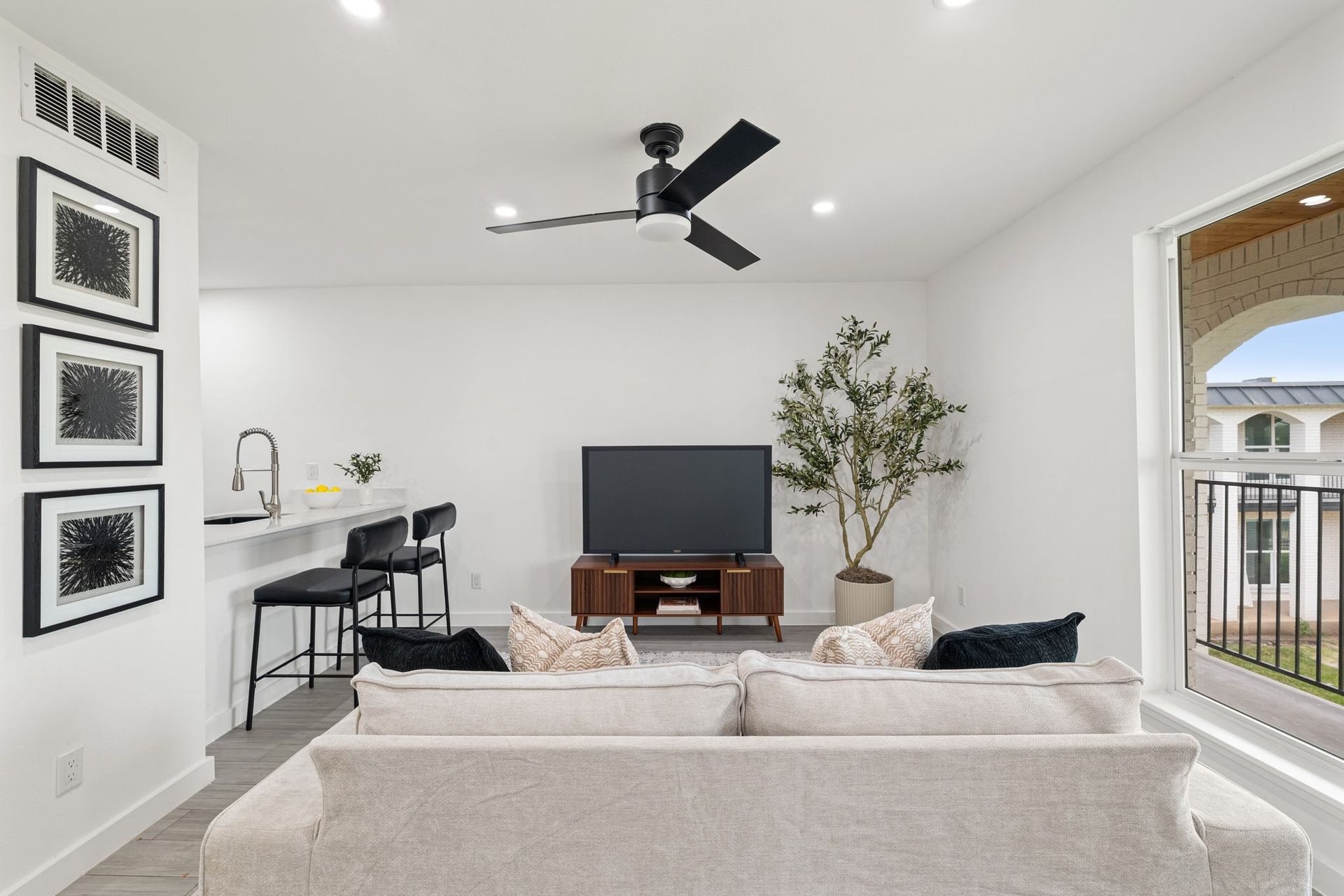 Bright living room with beige sofa, TV, bar stools, ceiling fan, and large windows overlooking a balcony
