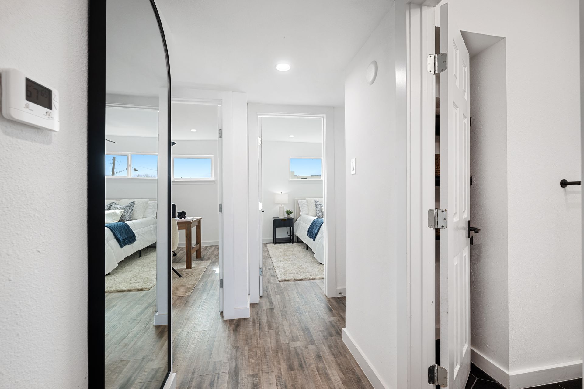 Bright hallway with white walls, wood floors, mirror closet doors, and a glimpse into a small bedroom.