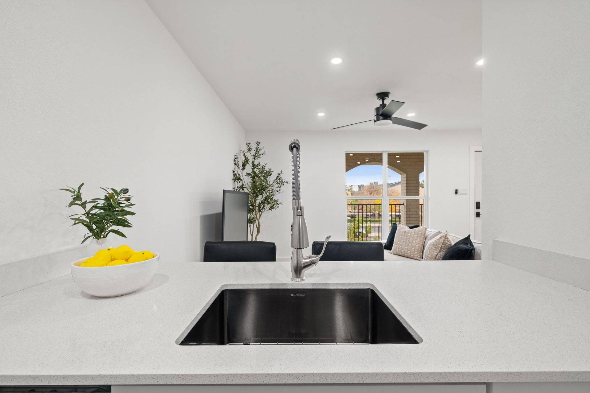 Bright modern kitchen with white countertops, black sink, and a view into a living room and patio.