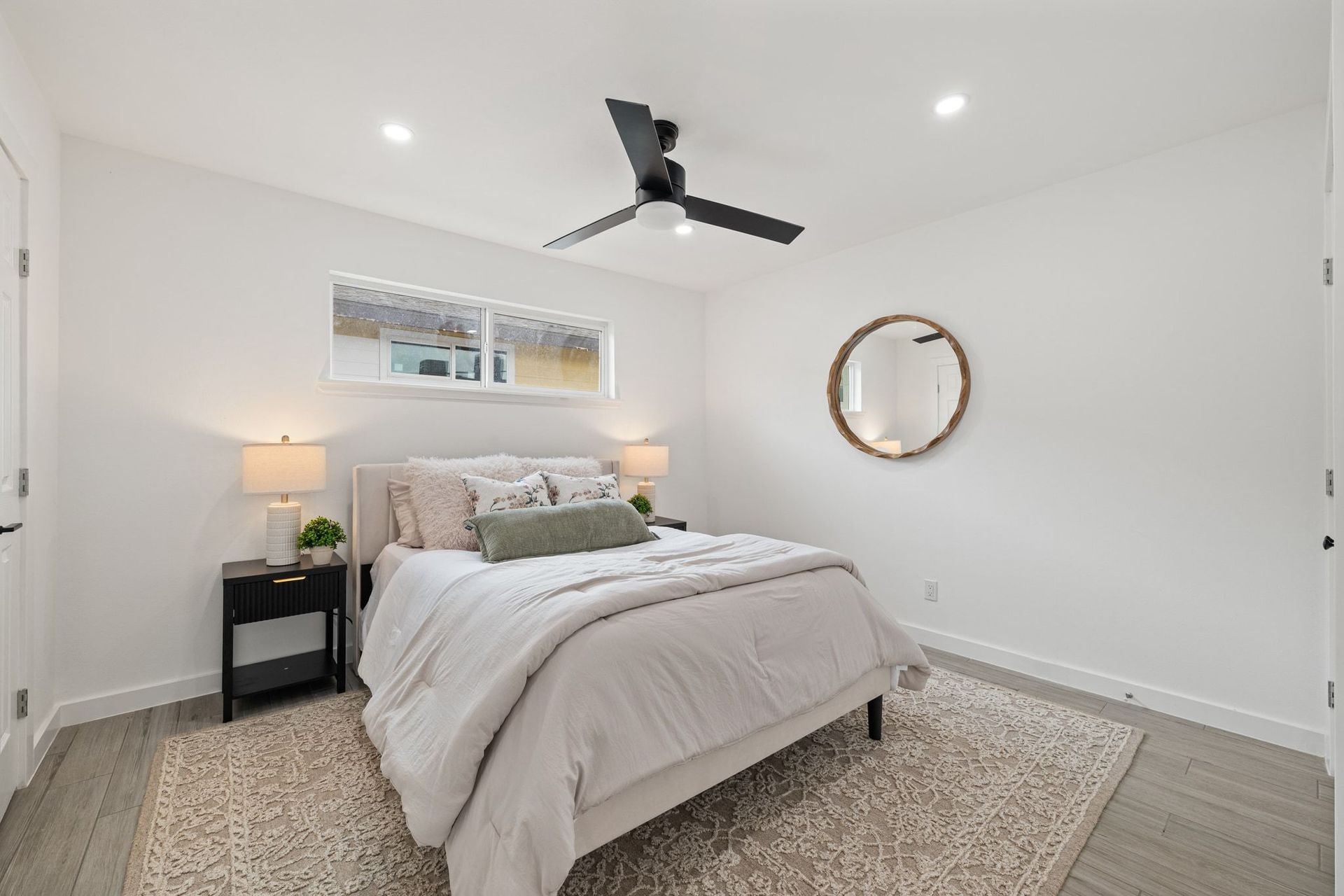 Bright modern bedroom with white walls, bed, nightstand, lamp, ceiling fan, and round mirror