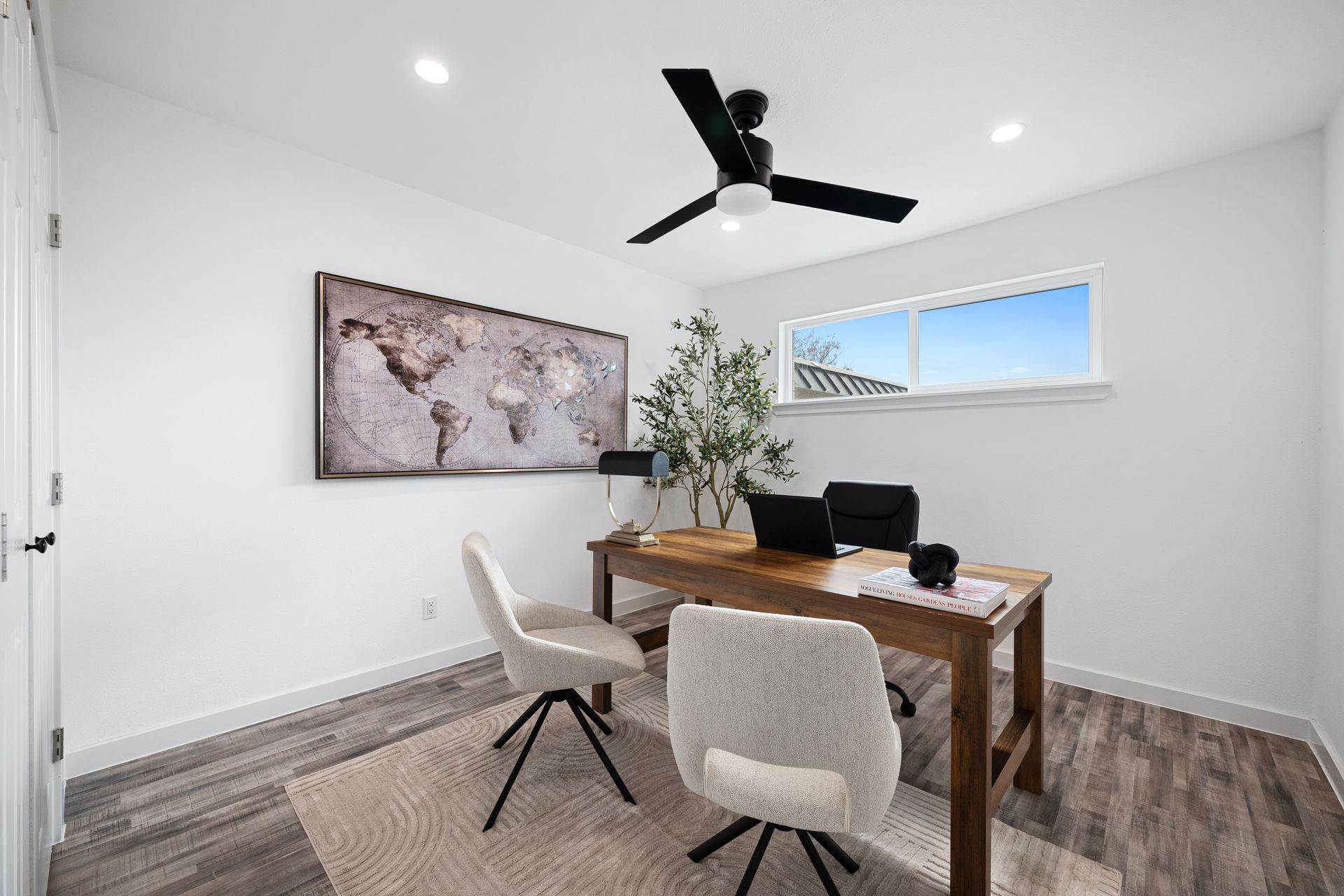 Bright home office with wooden desk, two chairs, wall map art, and a black ceiling fan.