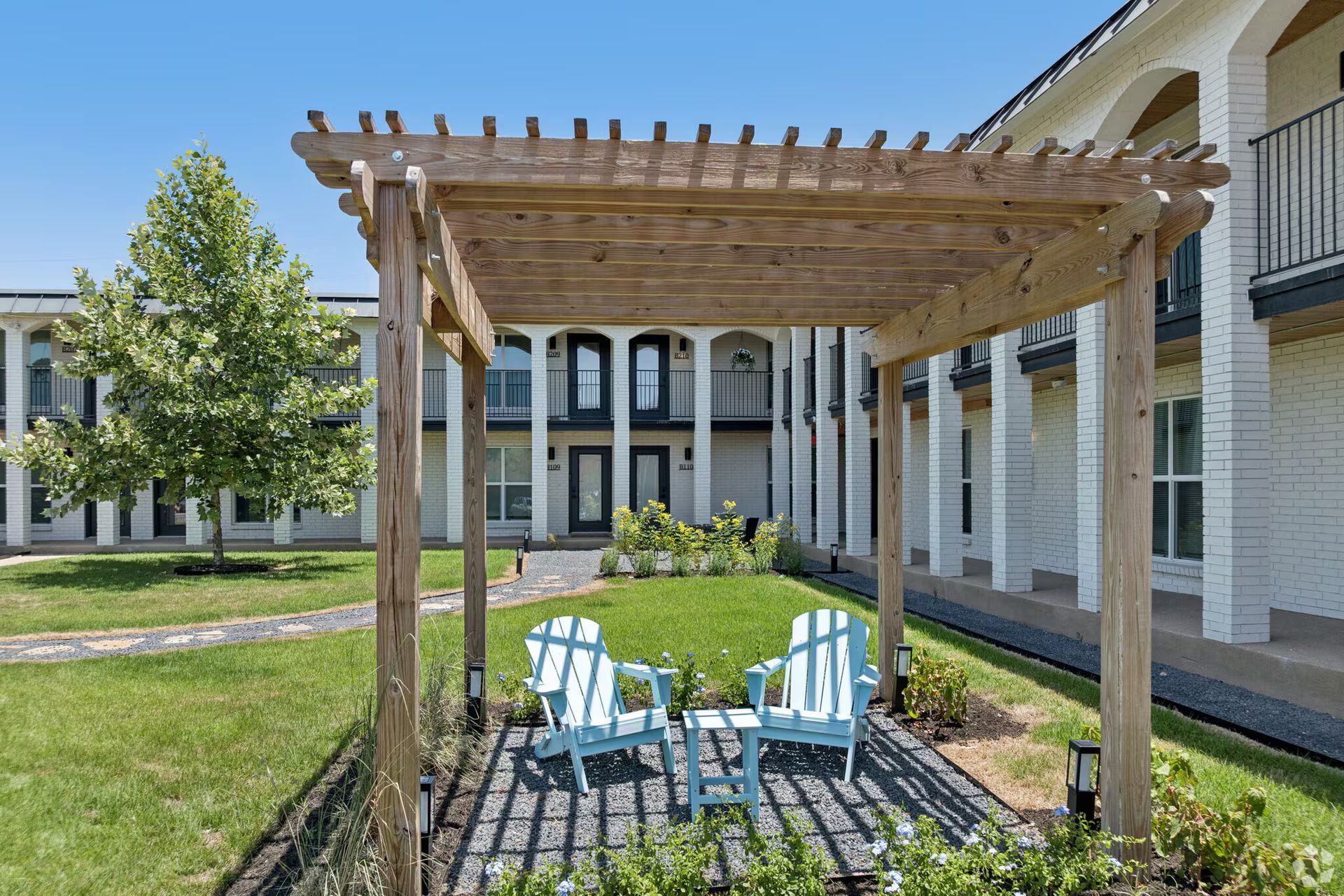 Wooden pergola with two blue chairs in a grassy courtyard between apartment buildings