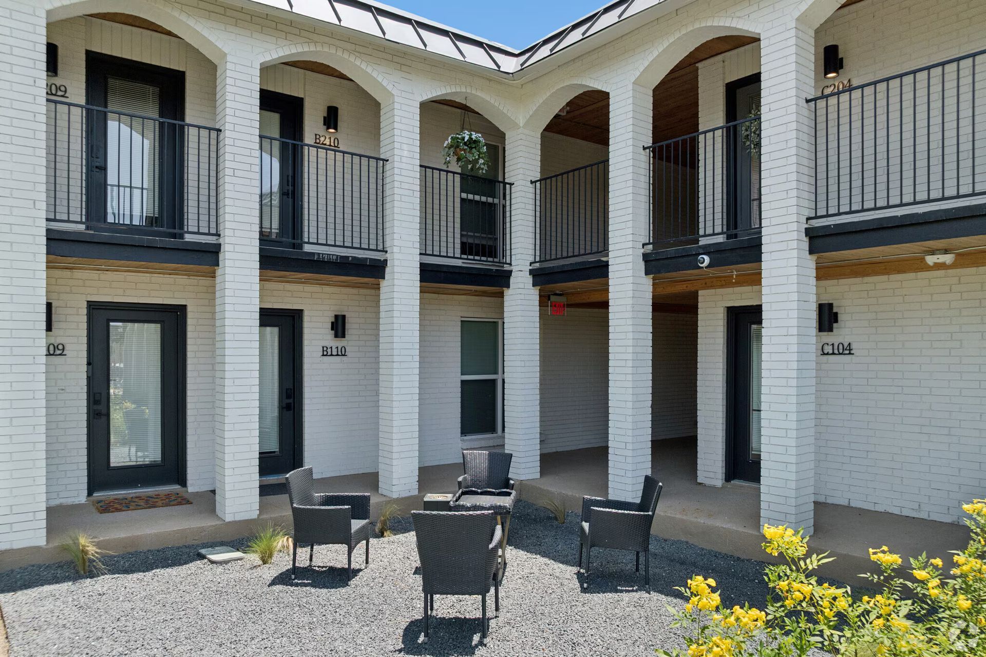 Courtyard of a two-story apartment building with black balconies, white walls, and outdoor chairs.