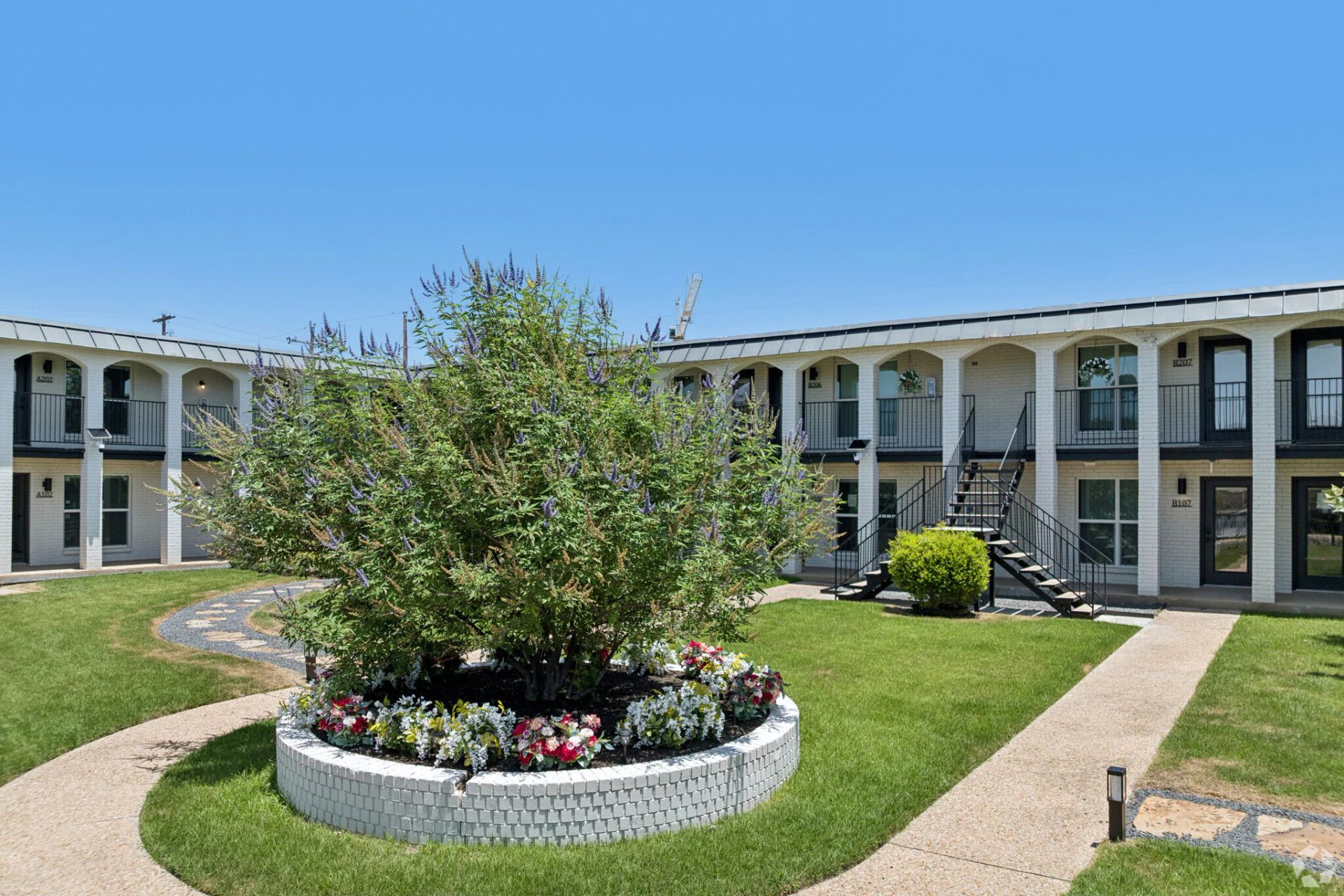 Courtyard with a flowering tree, circular flower bed, and two-story apartment buildings under a blue sky