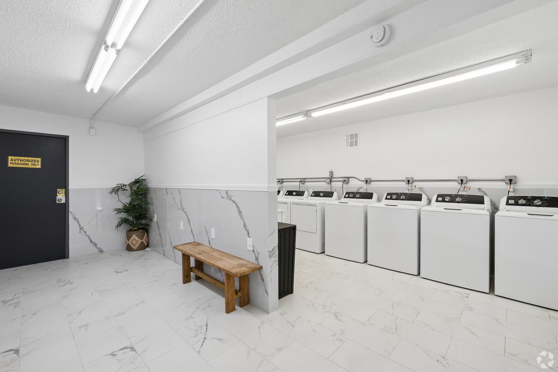 Bright laundry room with washers, white walls, a bench, and a potted plant