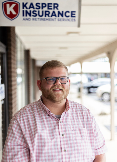 Man standing under a Kasper Insurance sign in a covered walkway, wearing a pink checkered shirt and glasses.