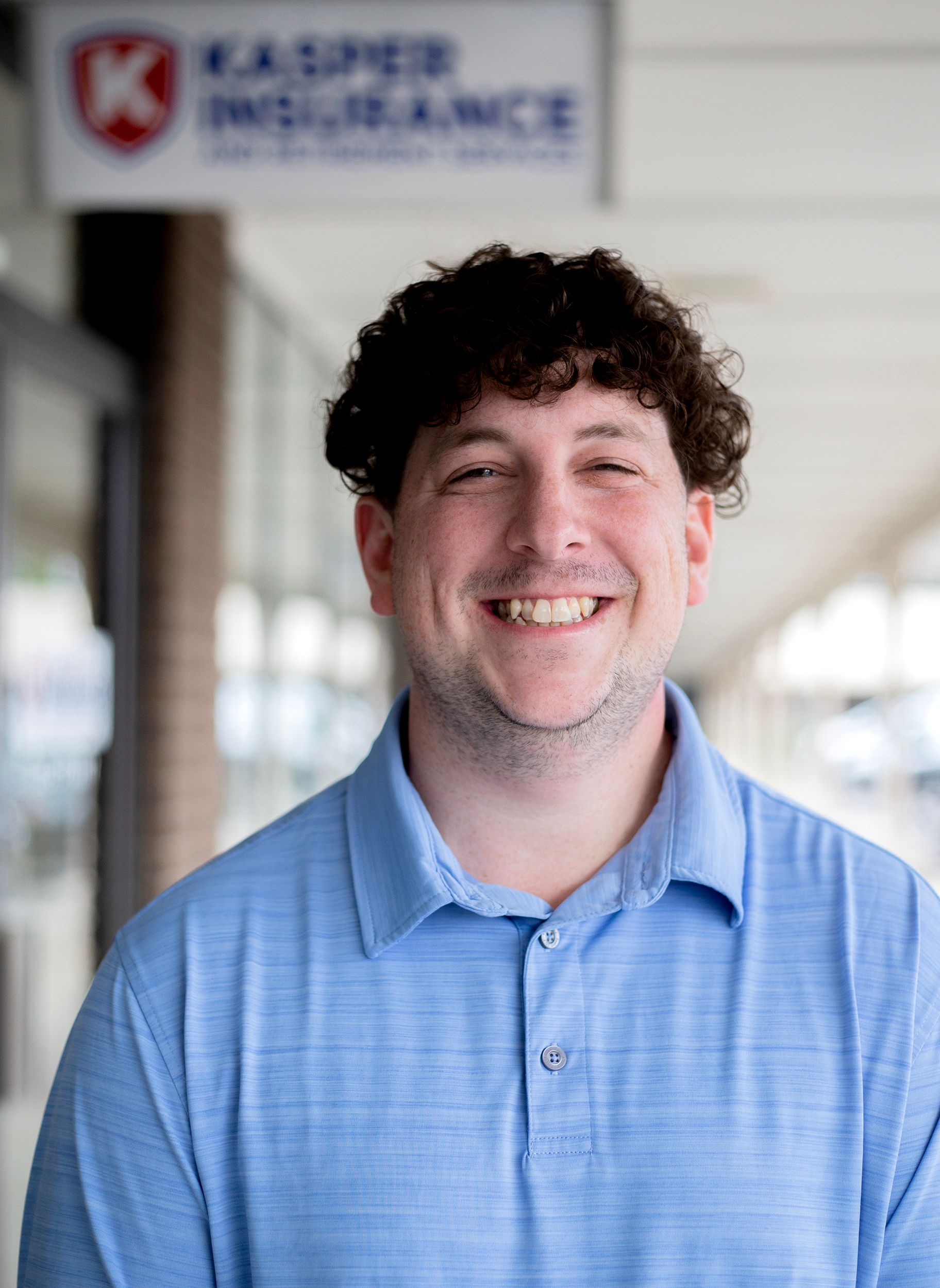 Smiling person in a light blue polo shirt standing outdoors in a blurred hallway