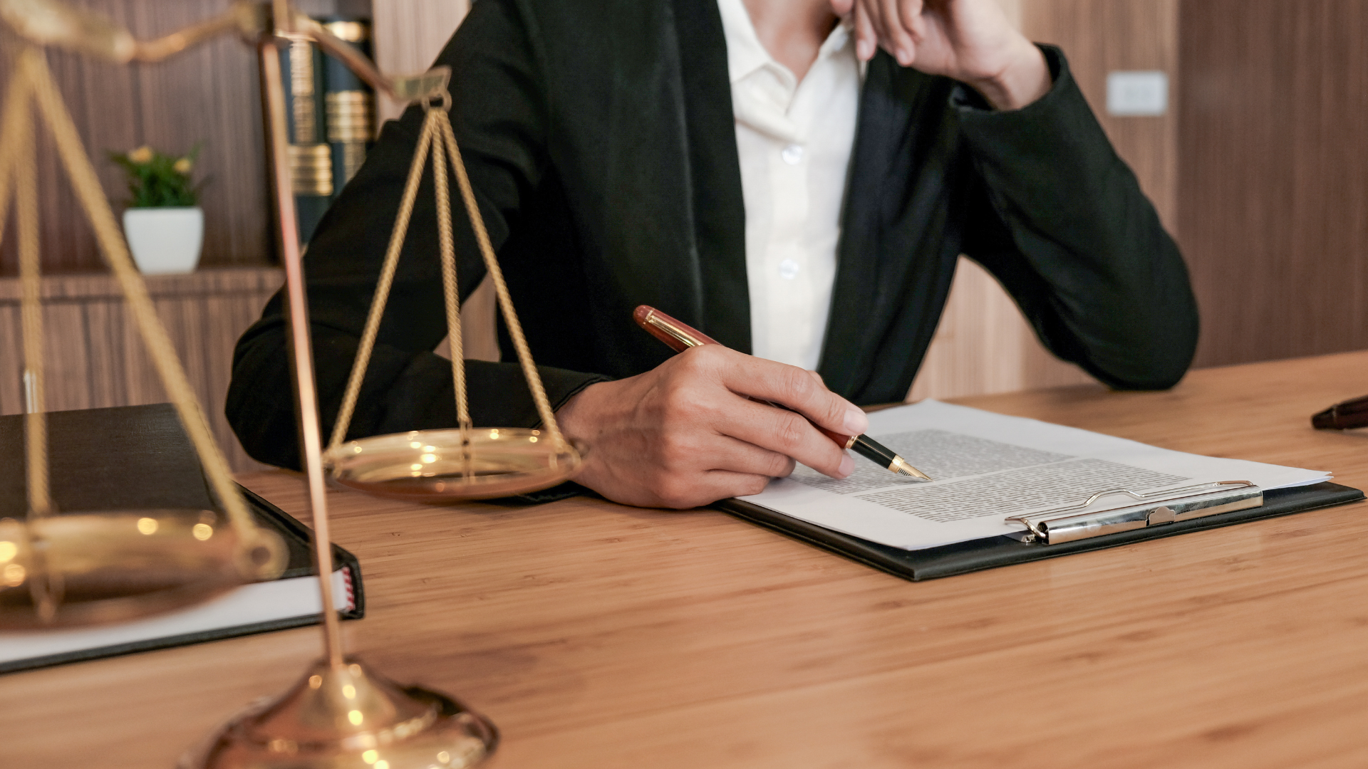 Person in a dark blazer writes in a notebook beside brass scales of justice on a wooden desk.