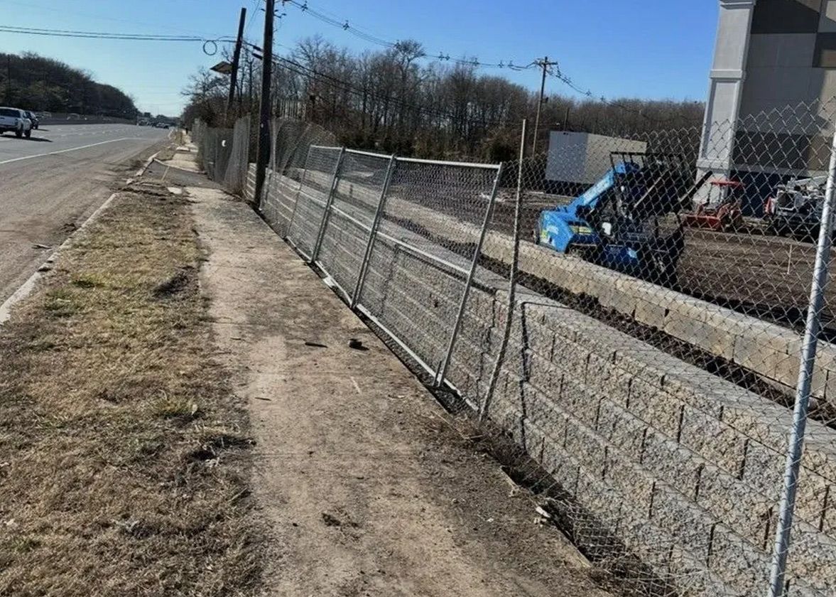 A sidewalk next to a road, separated by a low retaining wall with a chain-link fence and a gate.