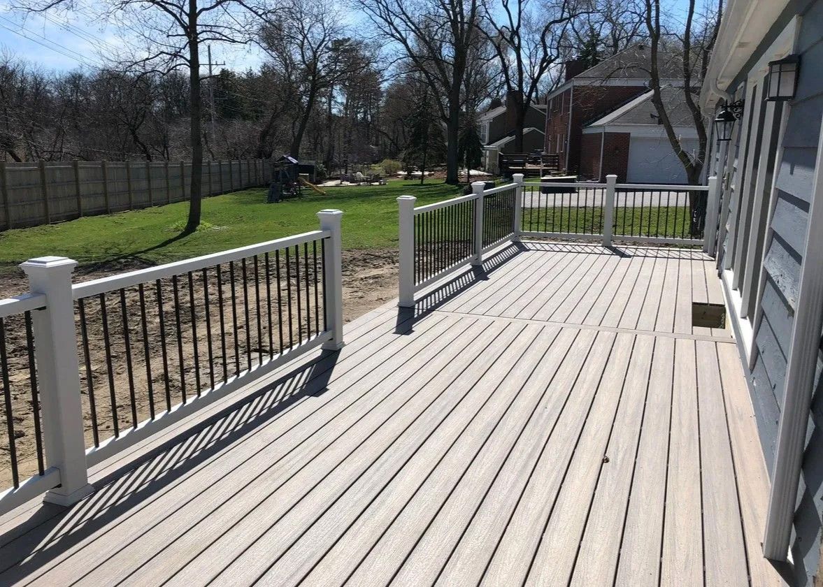 A wood deck with white posts and black spindles, next to a gray house, on a sunny day.