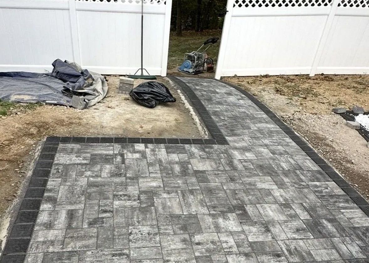 Paver pathway with black border leading to white fence gate. Construction materials lie nearby.