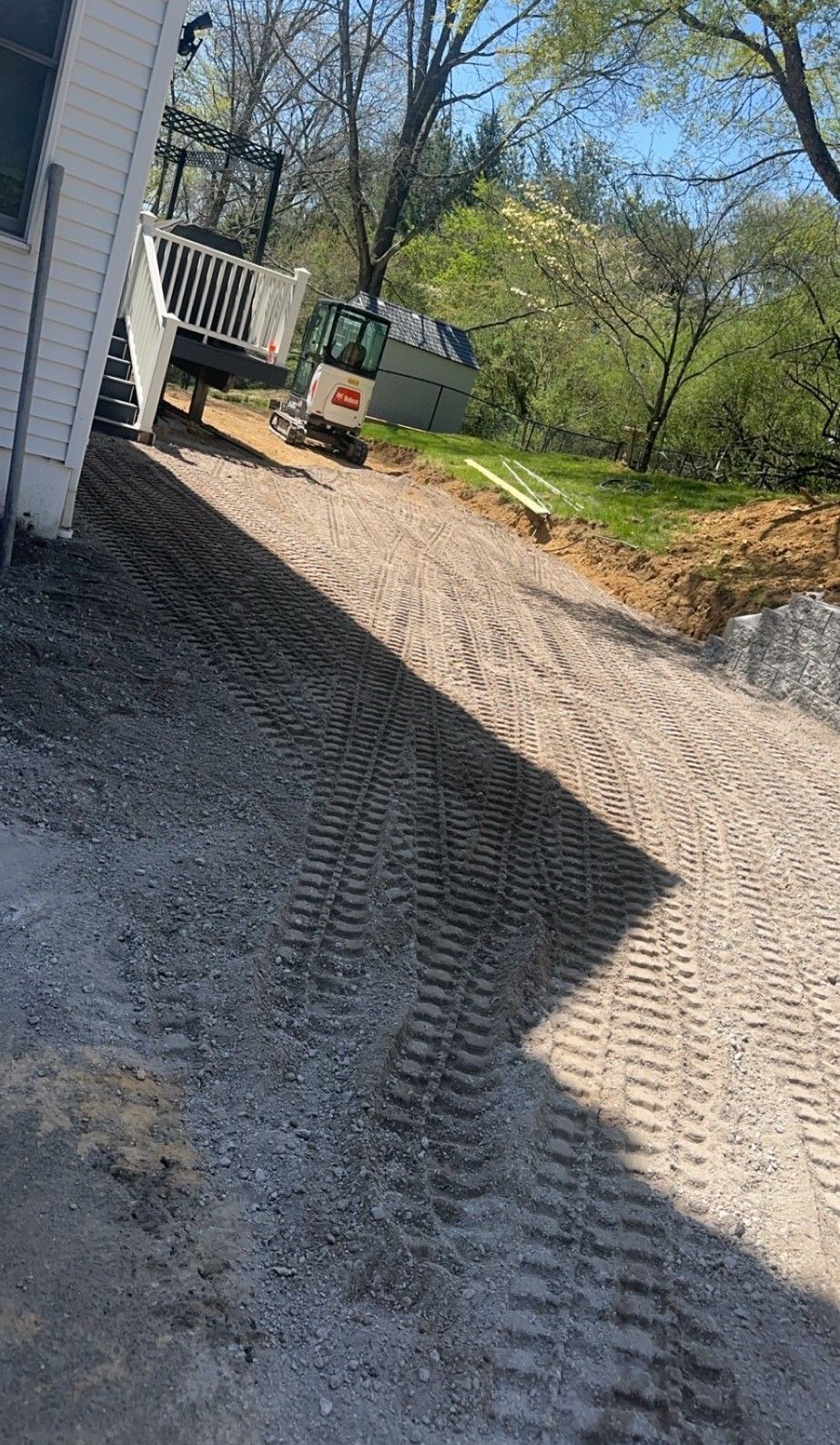 Gravel driveway leading up to a house with a small excavator parked near a deck on a sunny day.