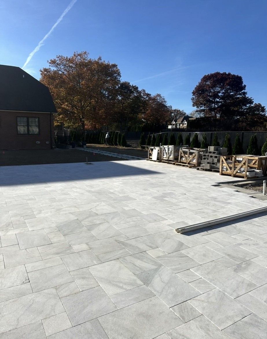 Light gray stone patio with building, trees, and construction materials on a sunny day.