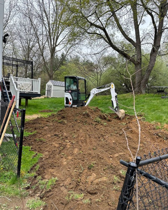 Mini excavator digging in a grassy yard next to a black fence and house.