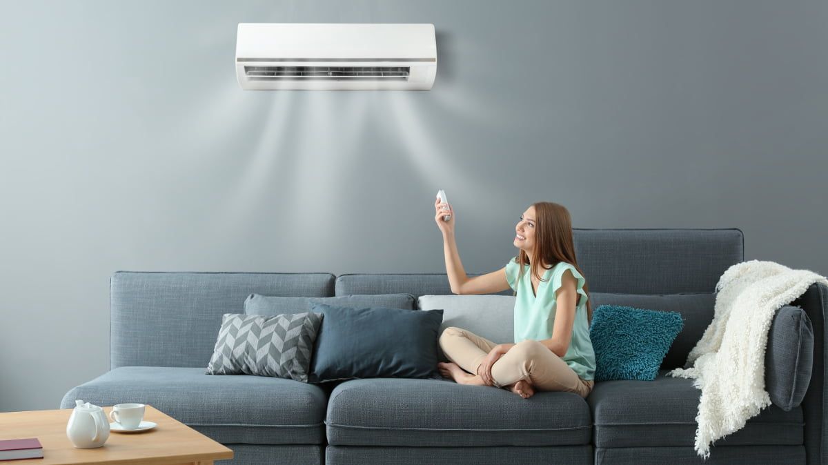 Mujer en sofá ajusta el aire acondicionado con control remoto, soplando aire fresco.