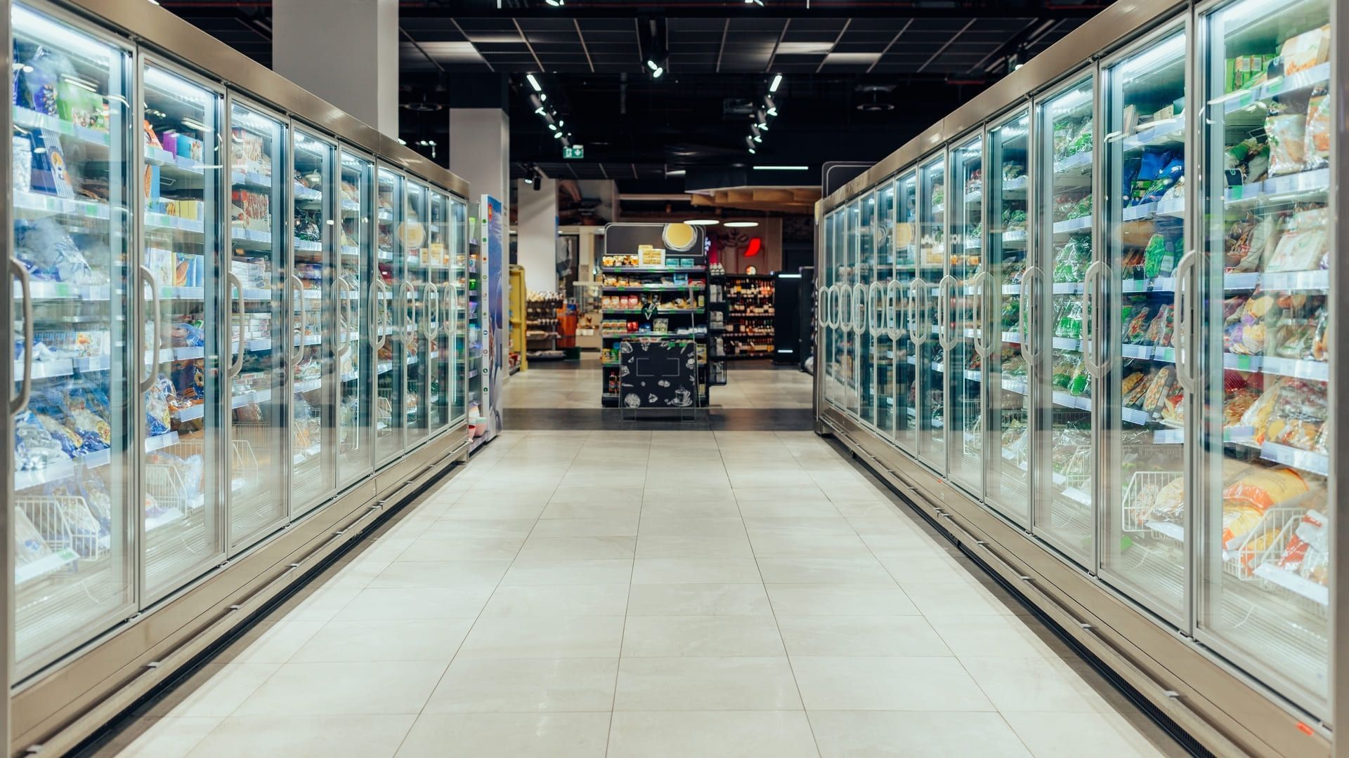 En un pasillo de un supermercado se alinean vitrinas de alimentos refrigerados, repletas de diversos productos.