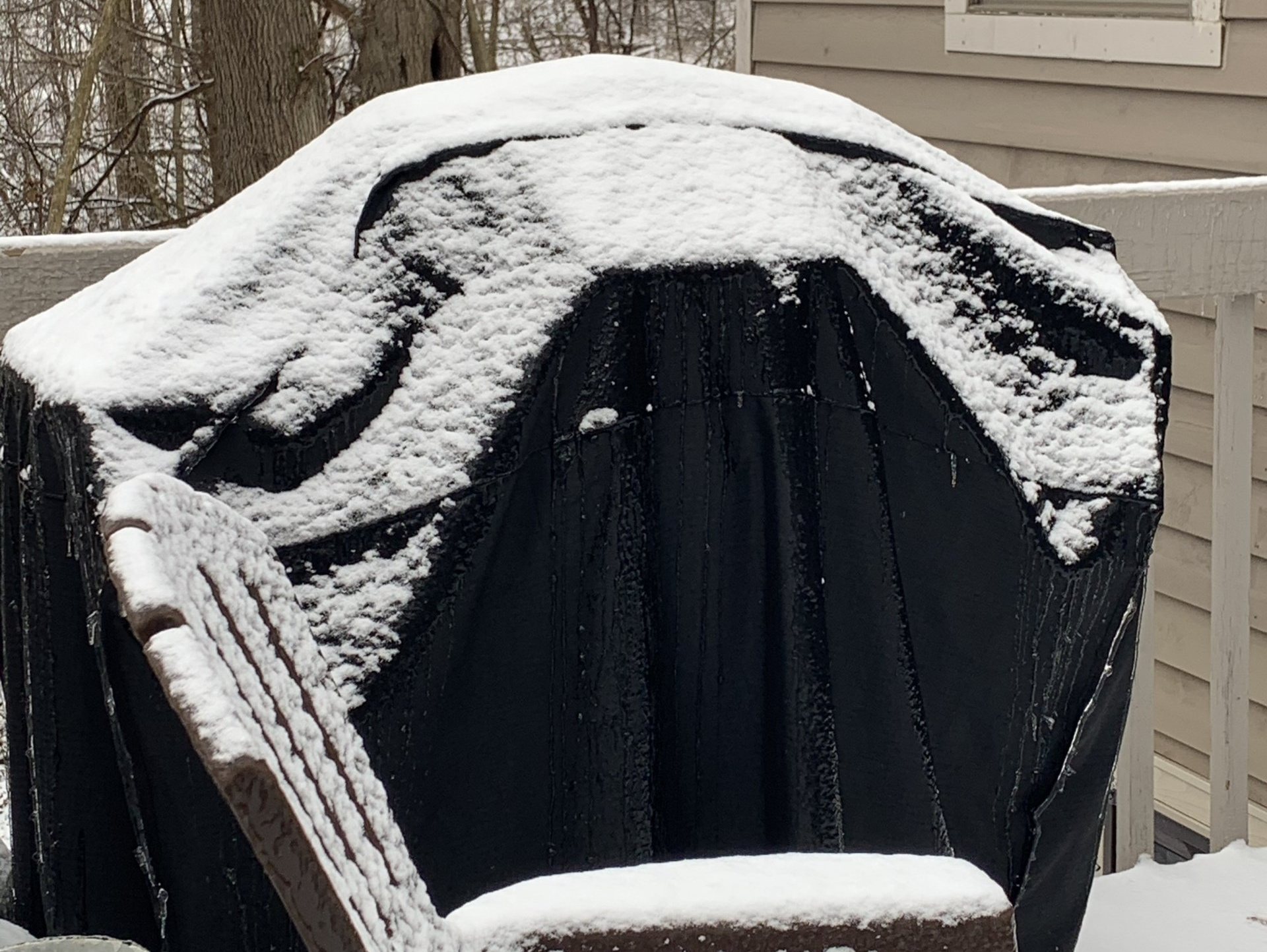 Snow-covered black grill cover on a wooden deck in winter.
