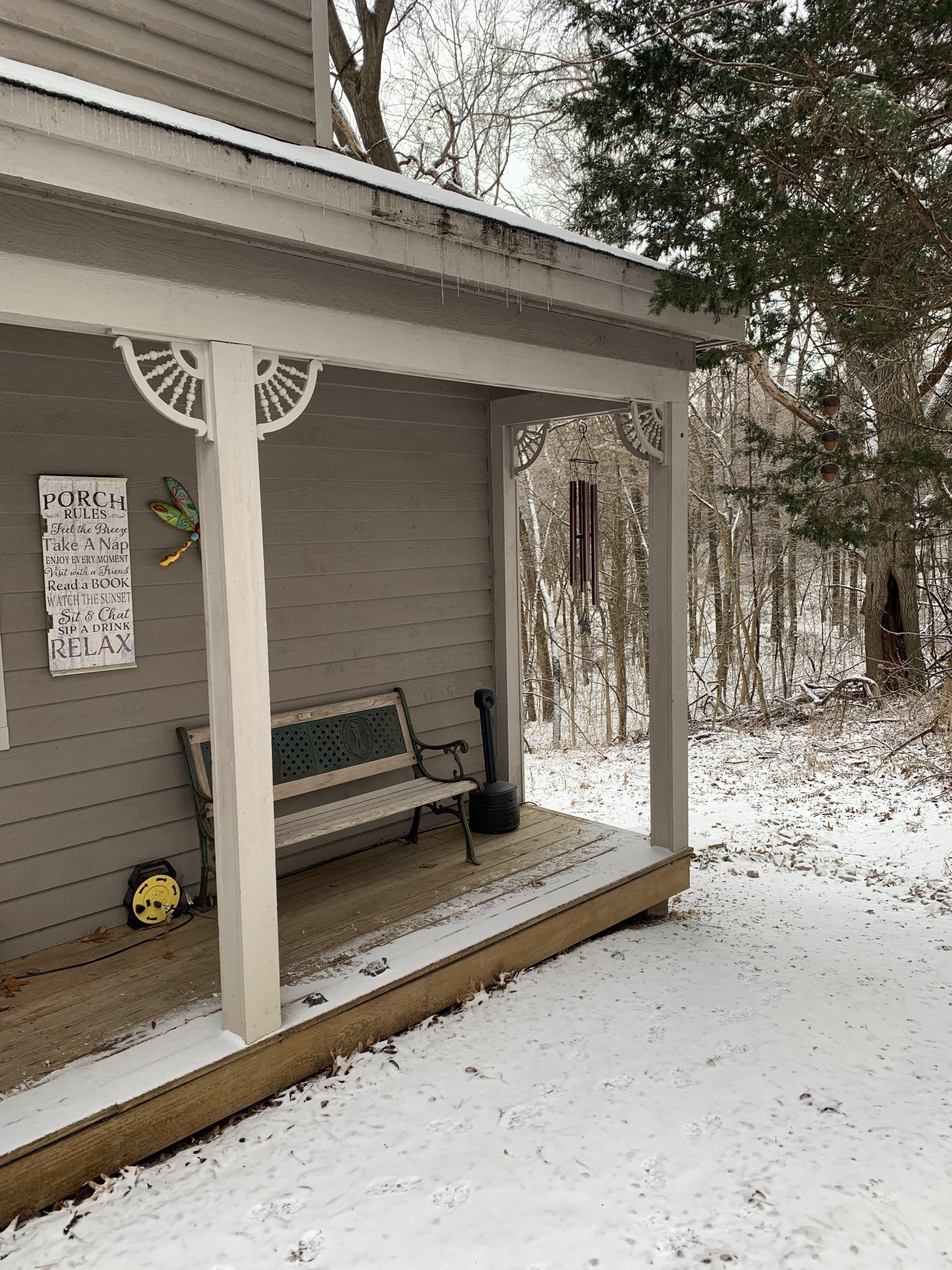Snowy porch with bench, decorative trim, and bird feeder. Trees in the background.