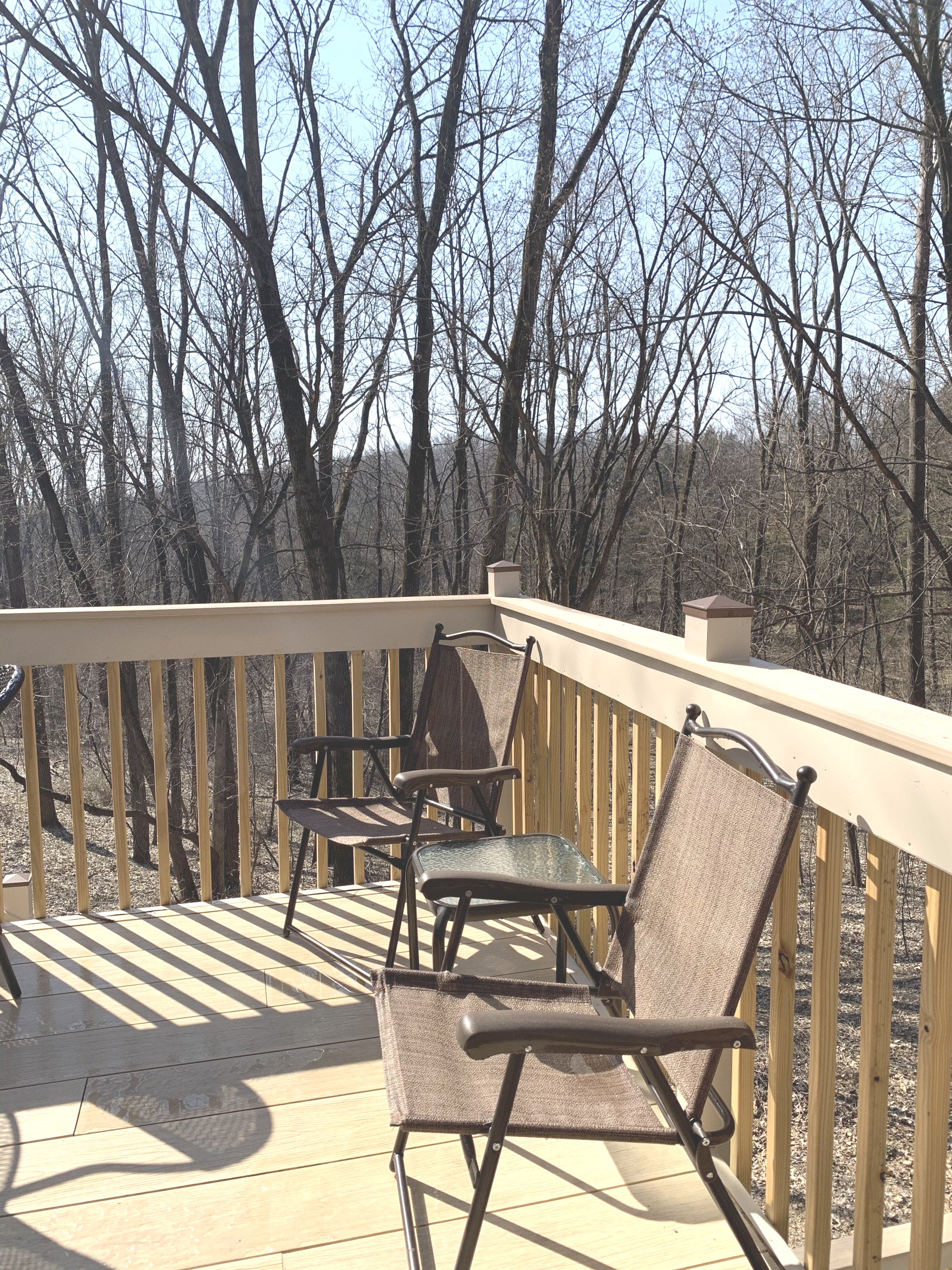 Wooden deck with two chairs and a table overlooking a bare tree forest on a sunny day.