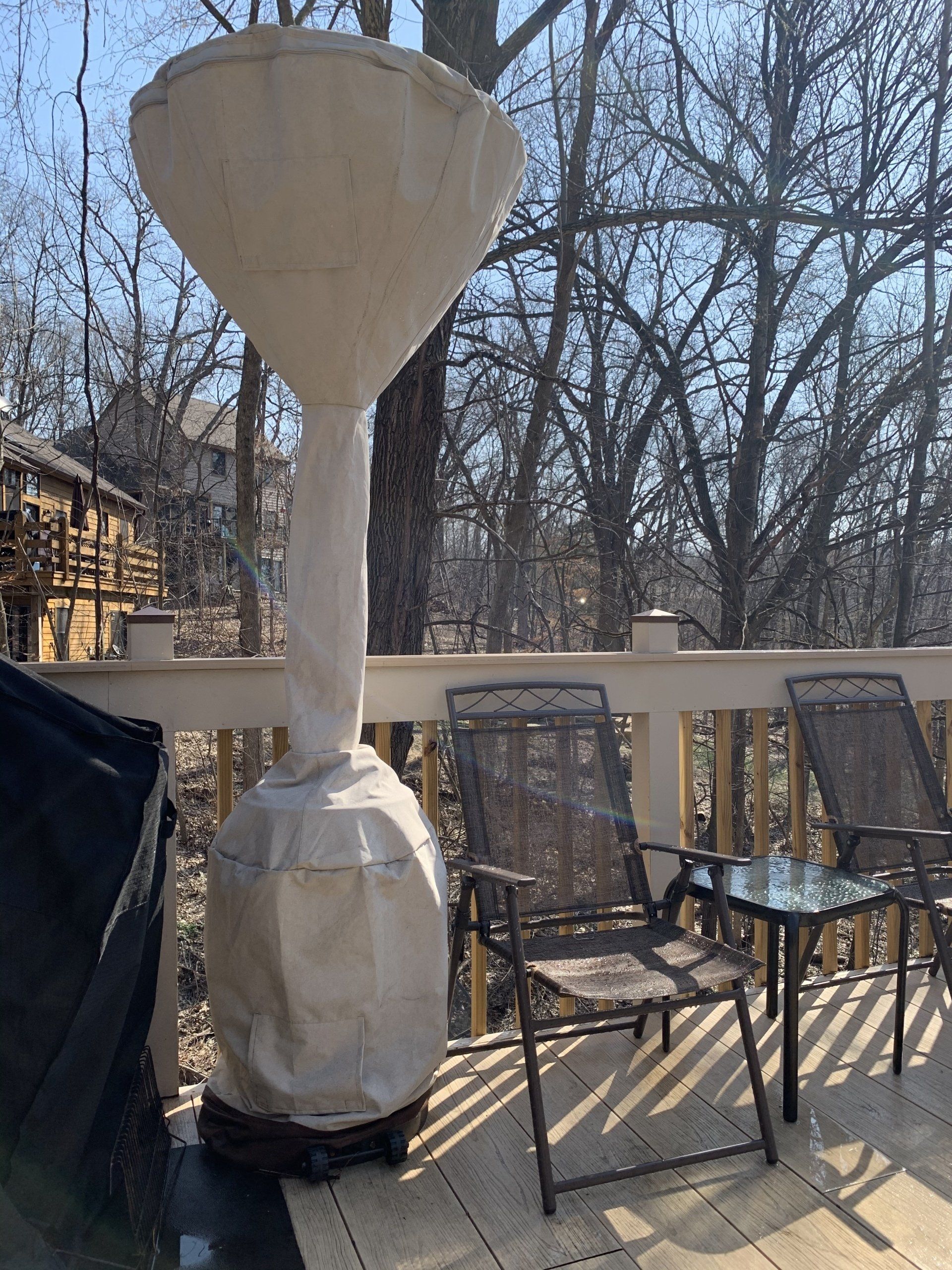 Patio heater with protective cover on a wooden deck with chairs and trees in background.