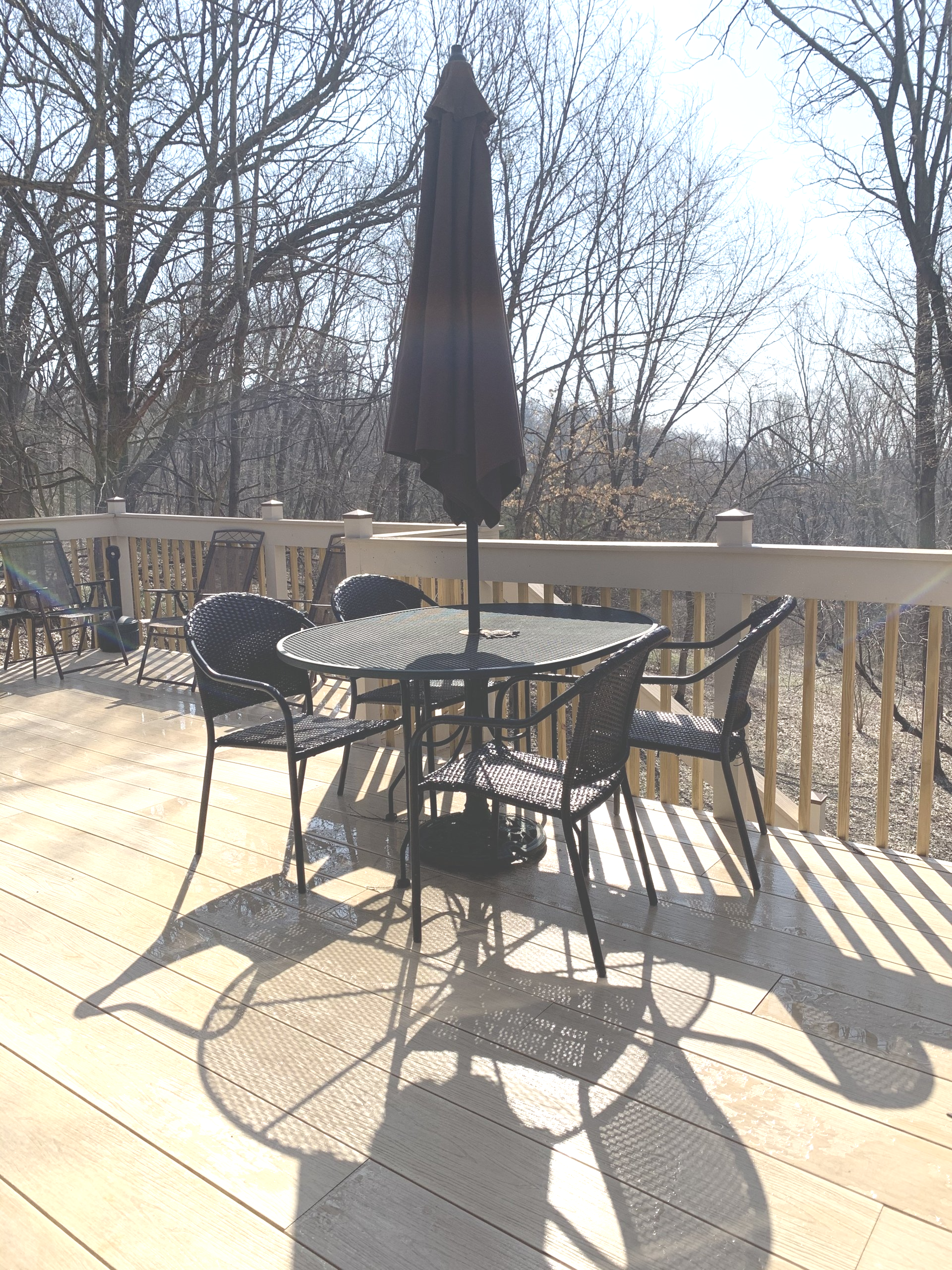 Patio set with table and chairs under an umbrella on a wooden deck in sunlight.