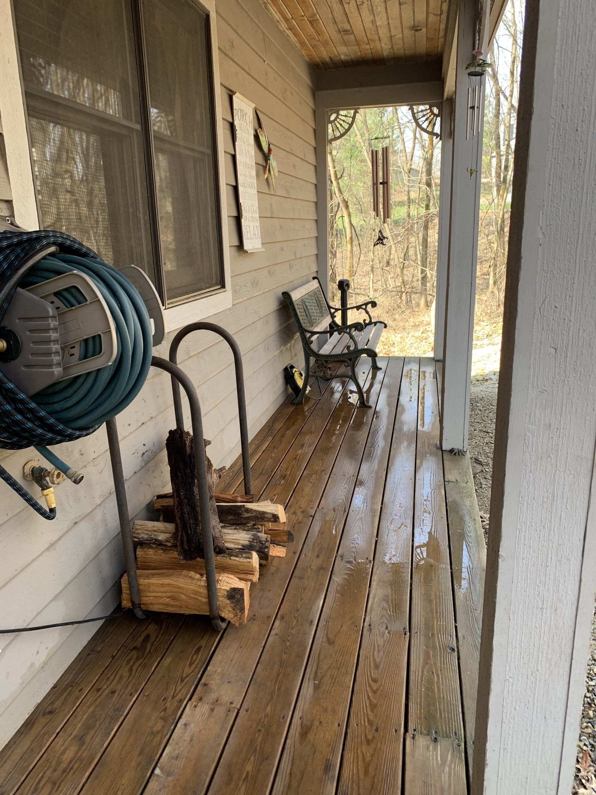 A covered porch with wet wood flooring, a bench, a hose, and a firewood rack.