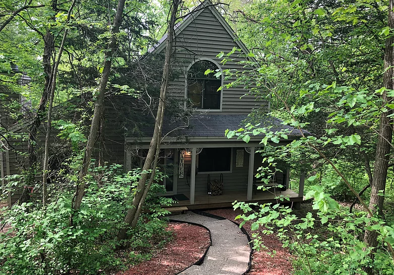 Two-story cabin nestled in trees. Stone walkway leads to porch. Grey siding, arched window, and green foliage.