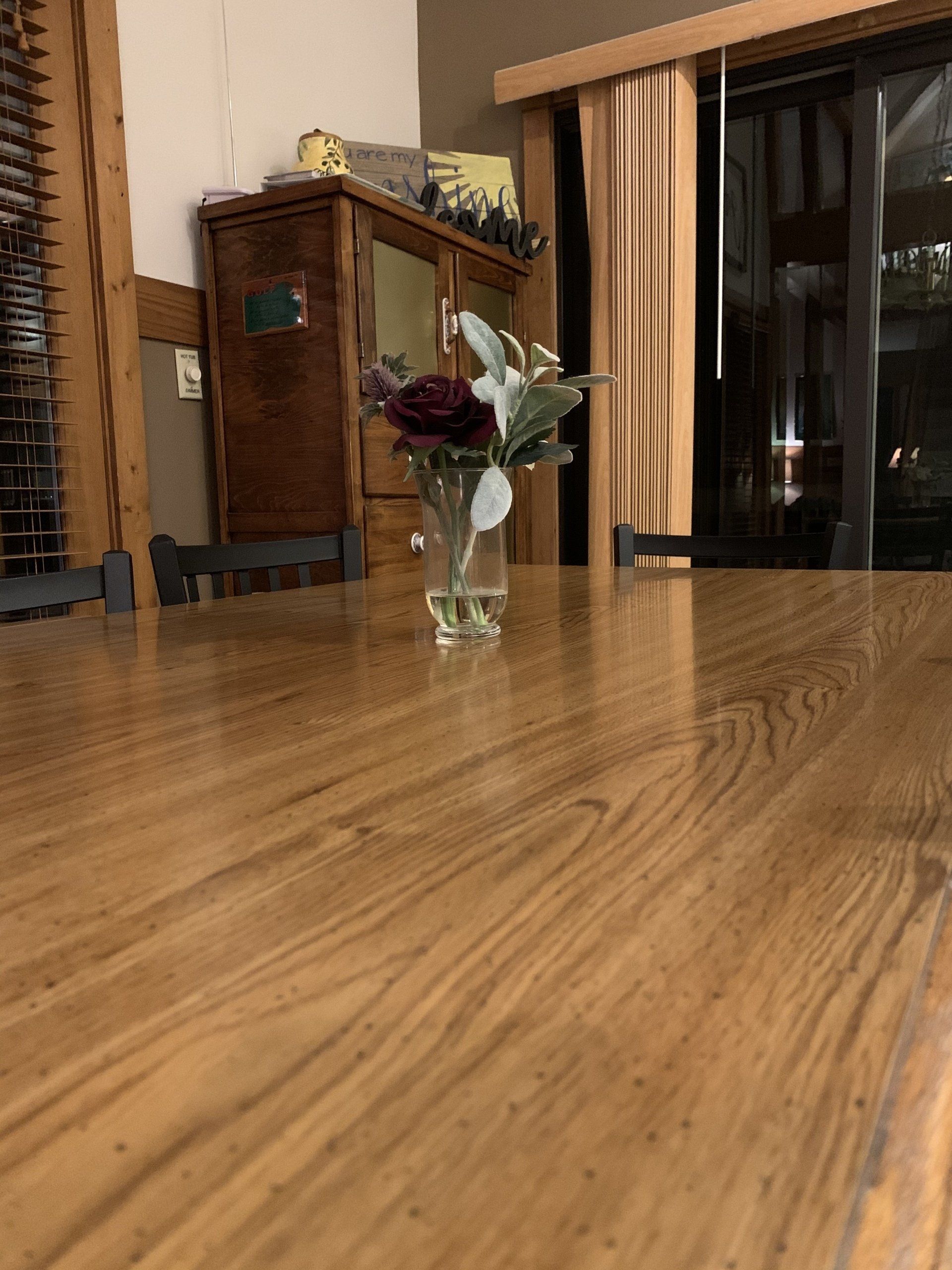Wooden dining table with a vase of flowers; a cabinet and window are visible in the background.