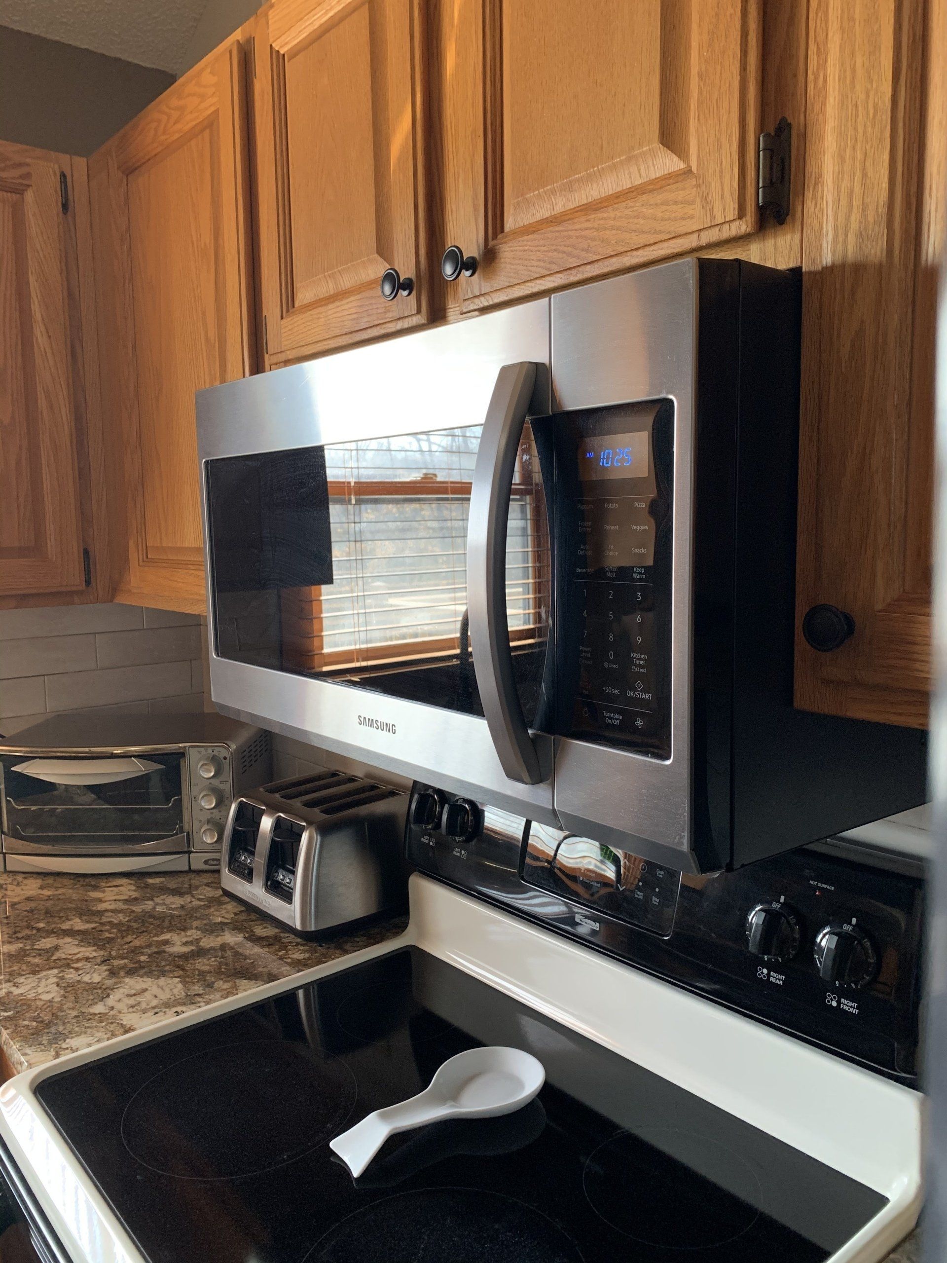 Stainless steel microwave over a stovetop in a kitchen. Wooden cabinets in background.