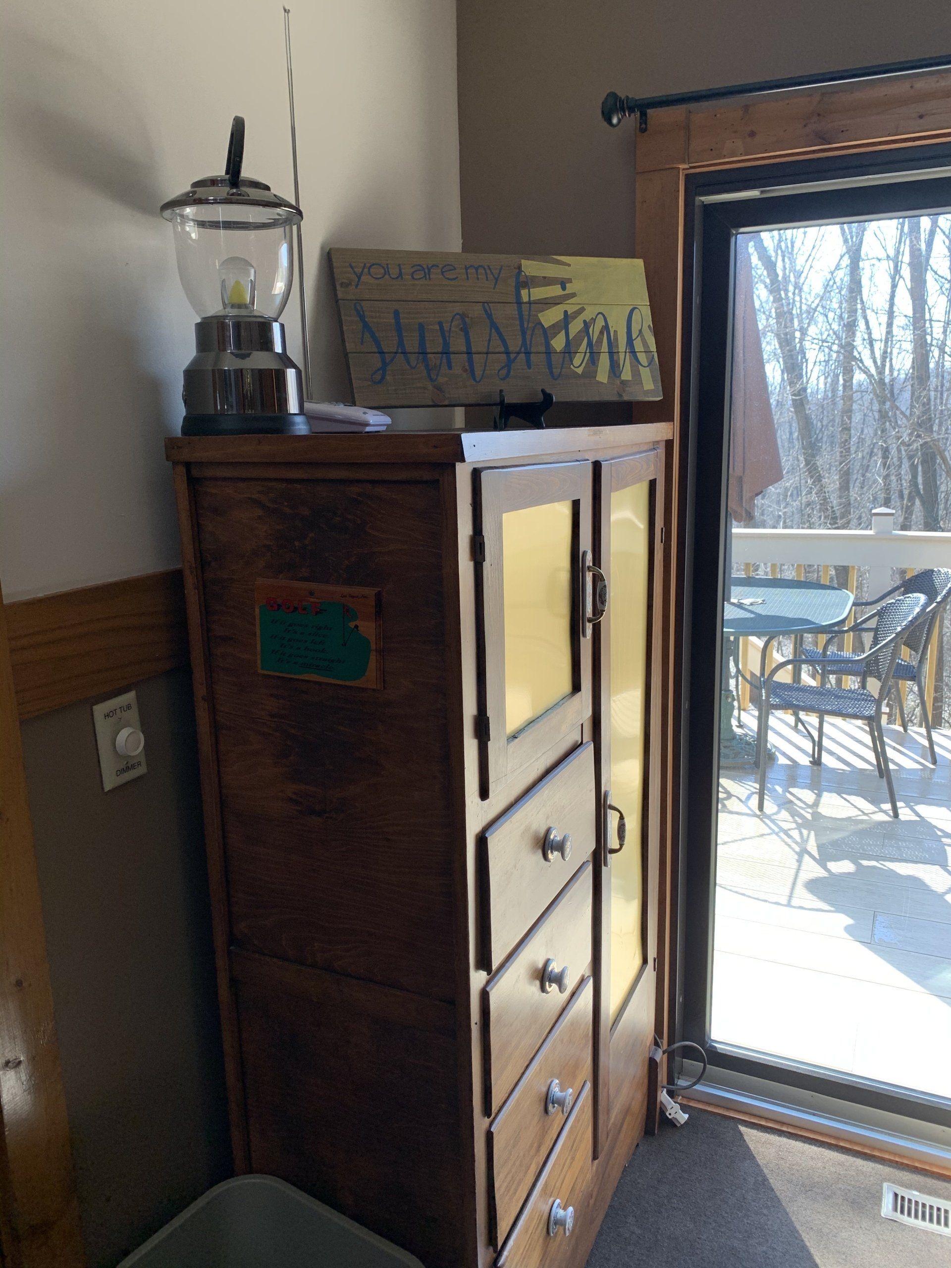 Tall wooden cabinet with drawers, a lantern, and a painting beside a door leading to a deck.