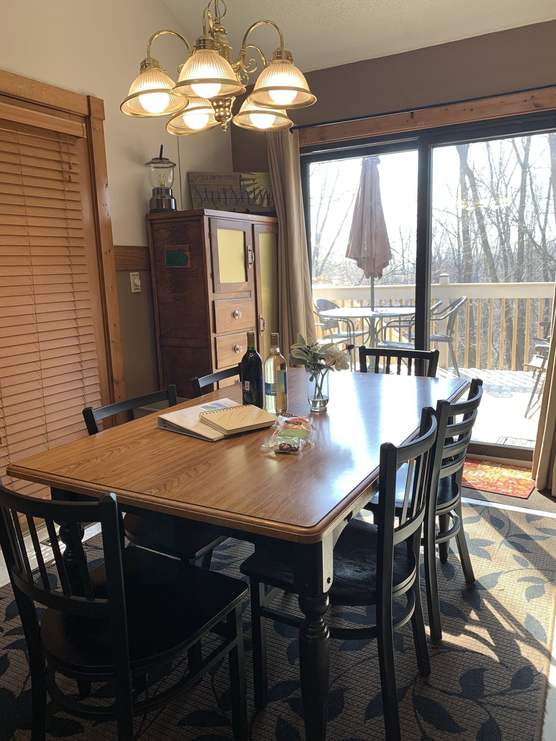 Dining room with wooden table, chairs, chandelier, and a view of a deck and trees.