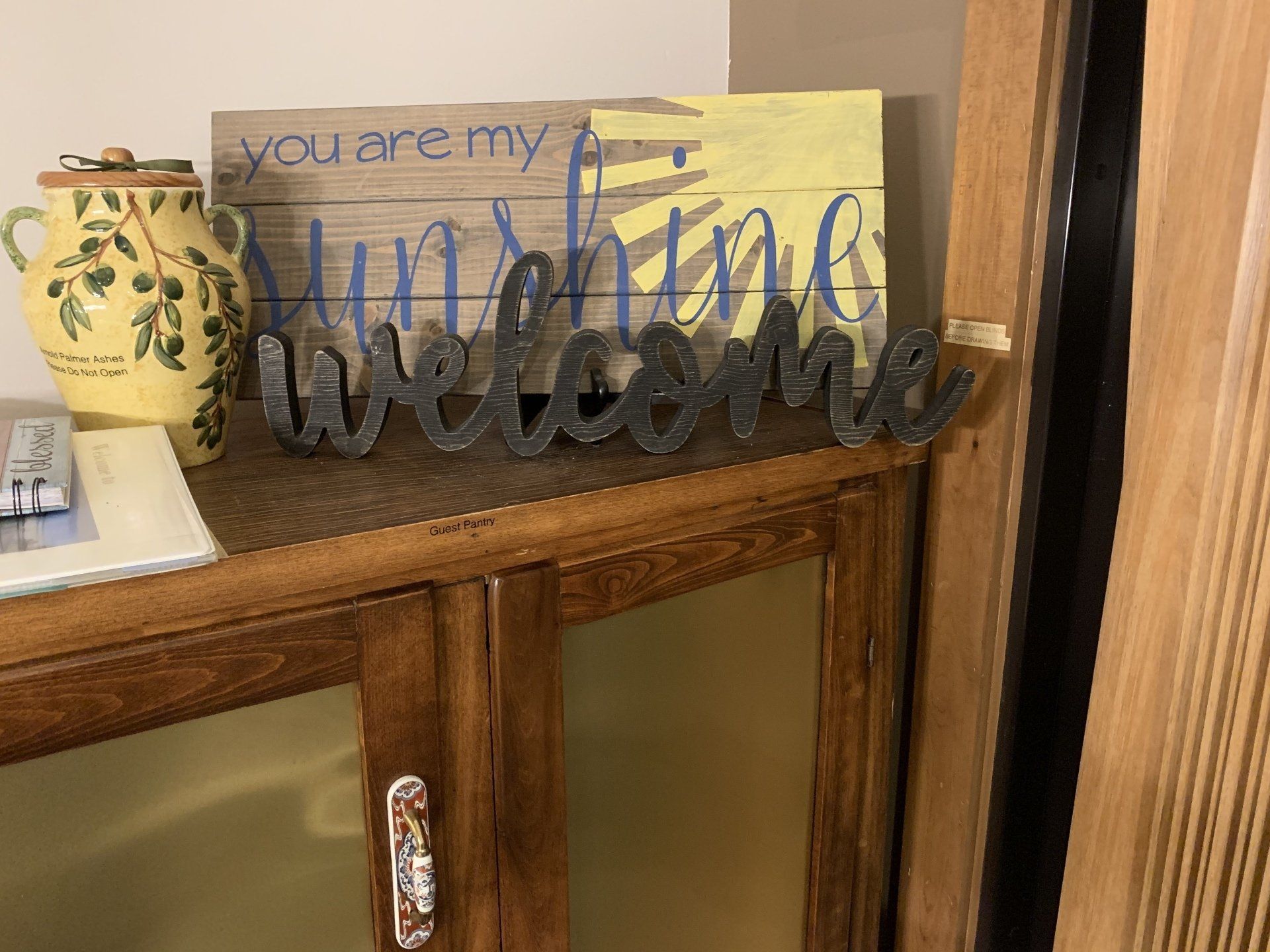 Wooden cabinet with welcome sign, sunshine art, and ceramic container.