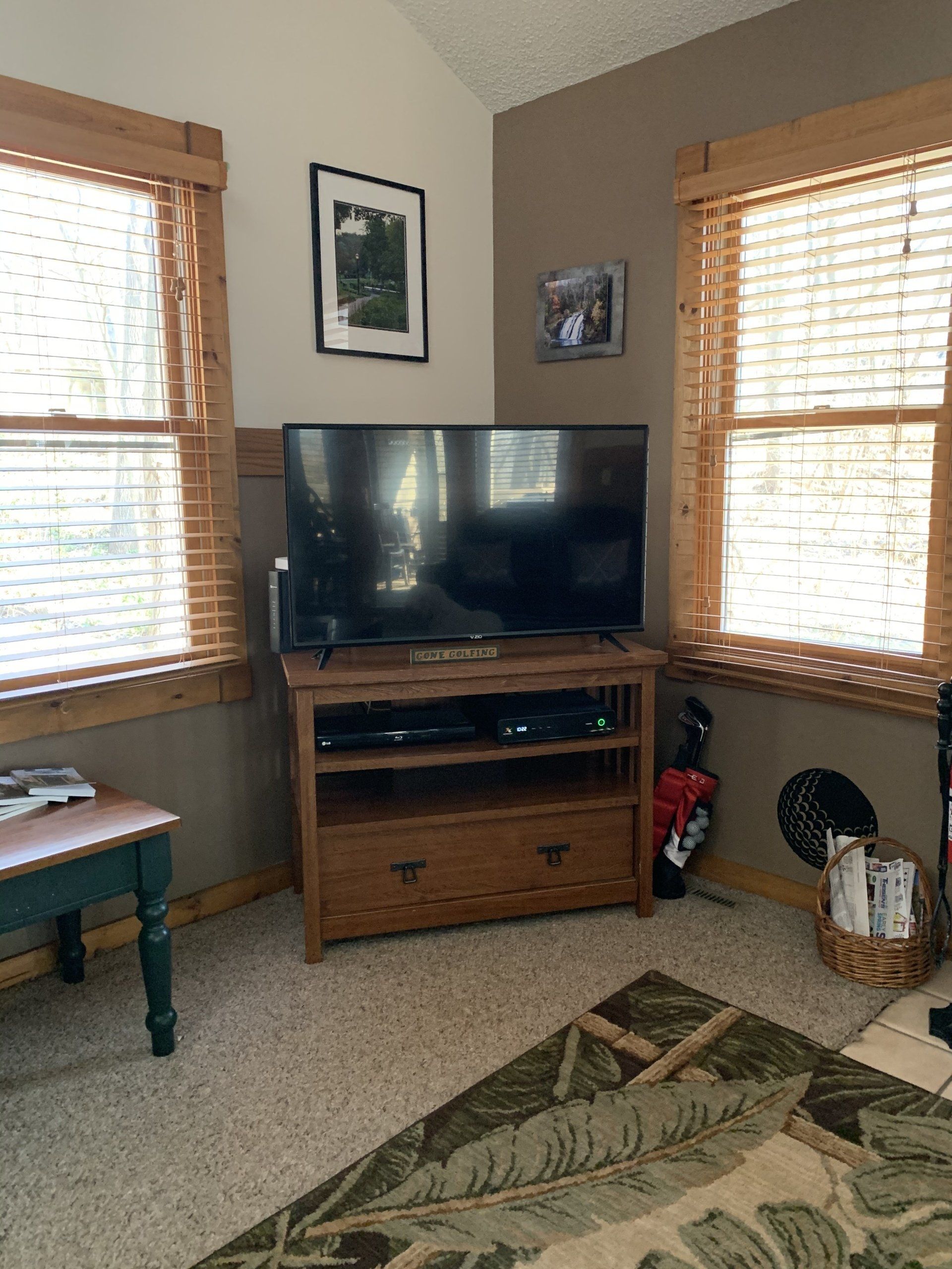 TV on a wooden cabinet between two windows with blinds; small bench and rug visible.
