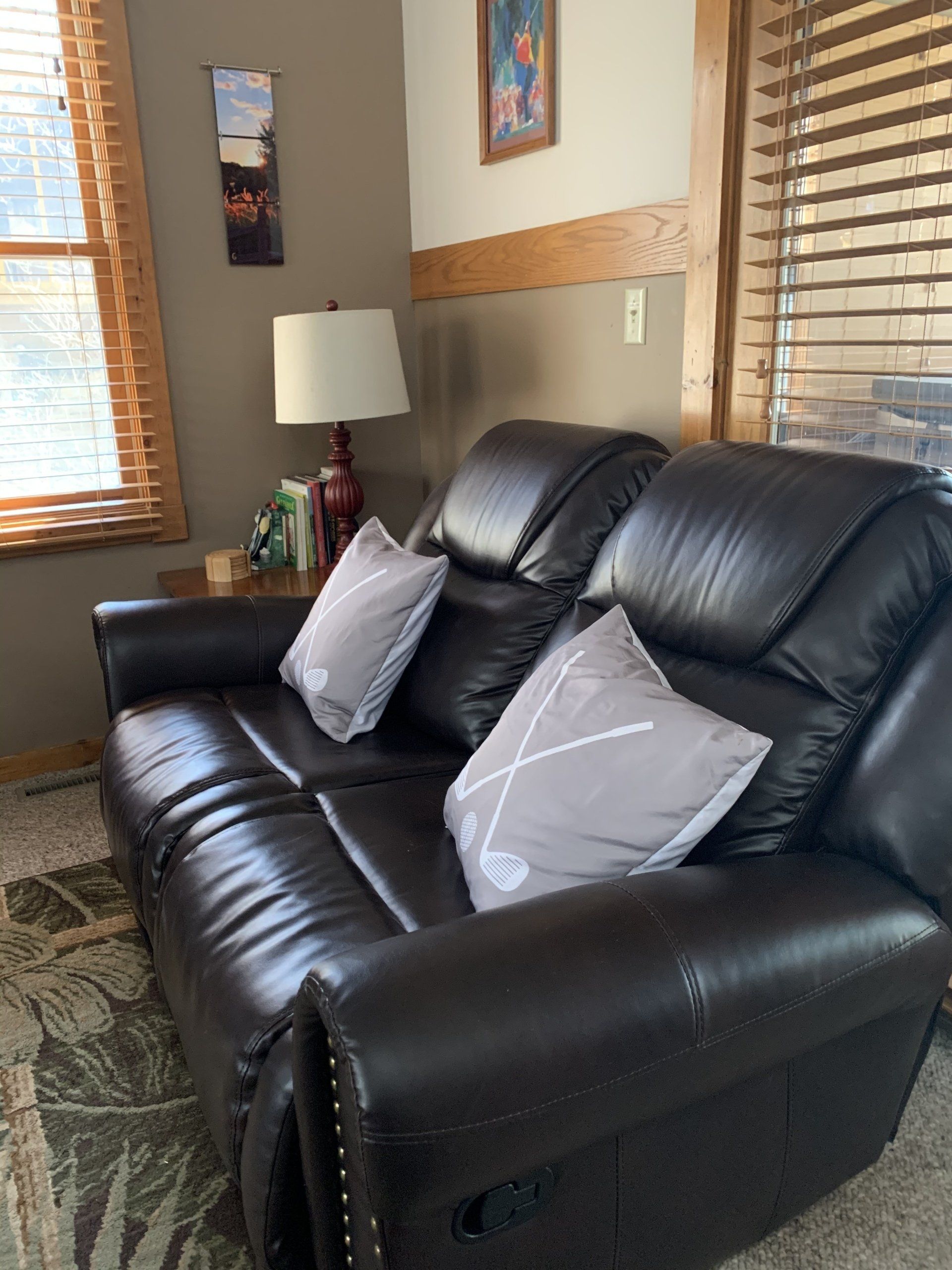 Dark brown leather loveseat with two gray pillows, beside a window with blinds, and a side table with lamp.