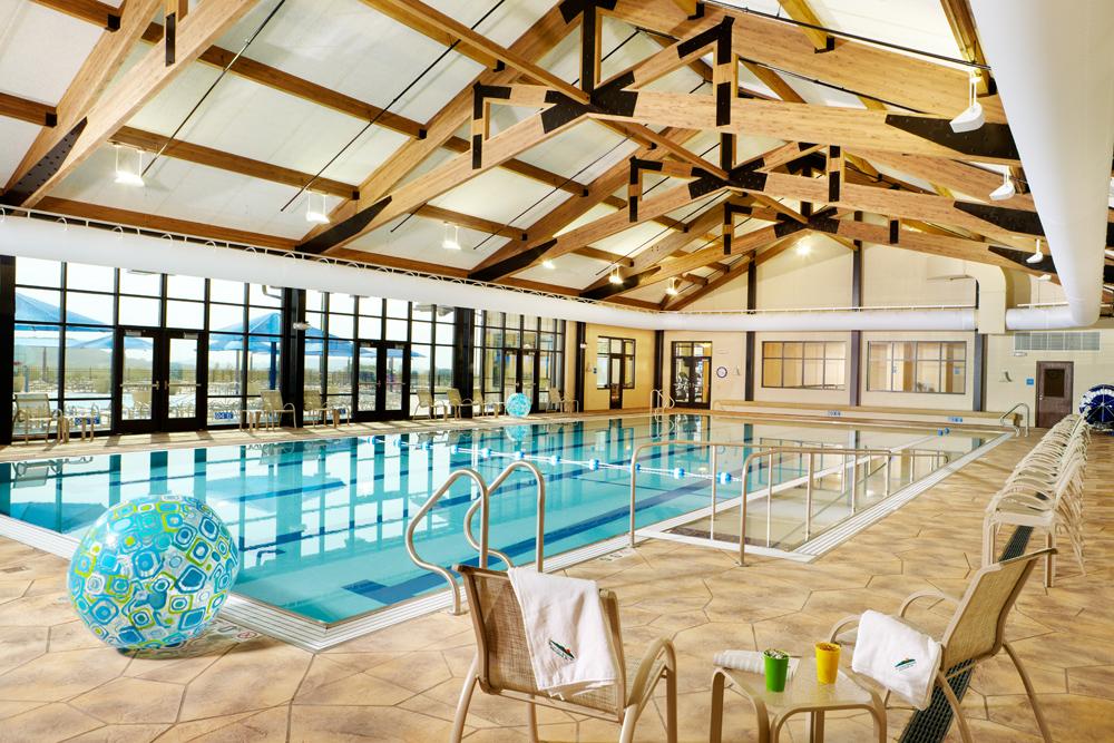 Indoor swimming pool with blue water, large windows, wooden beams, lounge chairs, and an inflatable ball.