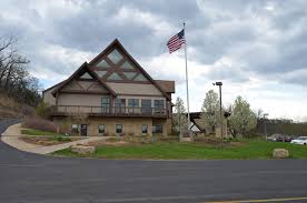 Two-story tan building with a triangular roof, an American flag, and a paved circular drive.