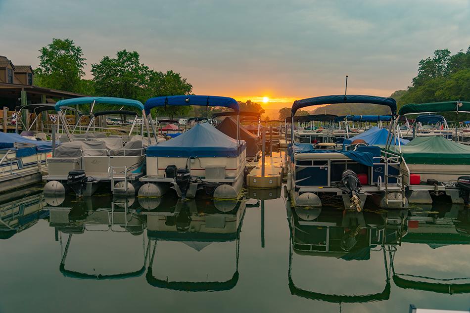 Boats docked in a harbor at sunset. Reflective water with sun and clouds overhead.