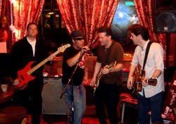 Band performing live in a dimly lit venue. Four men play instruments: electric guitar, bass, and drums.