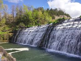 Waterfall cascading down rock tiers into a dark pool, with trees and blue sky visible.