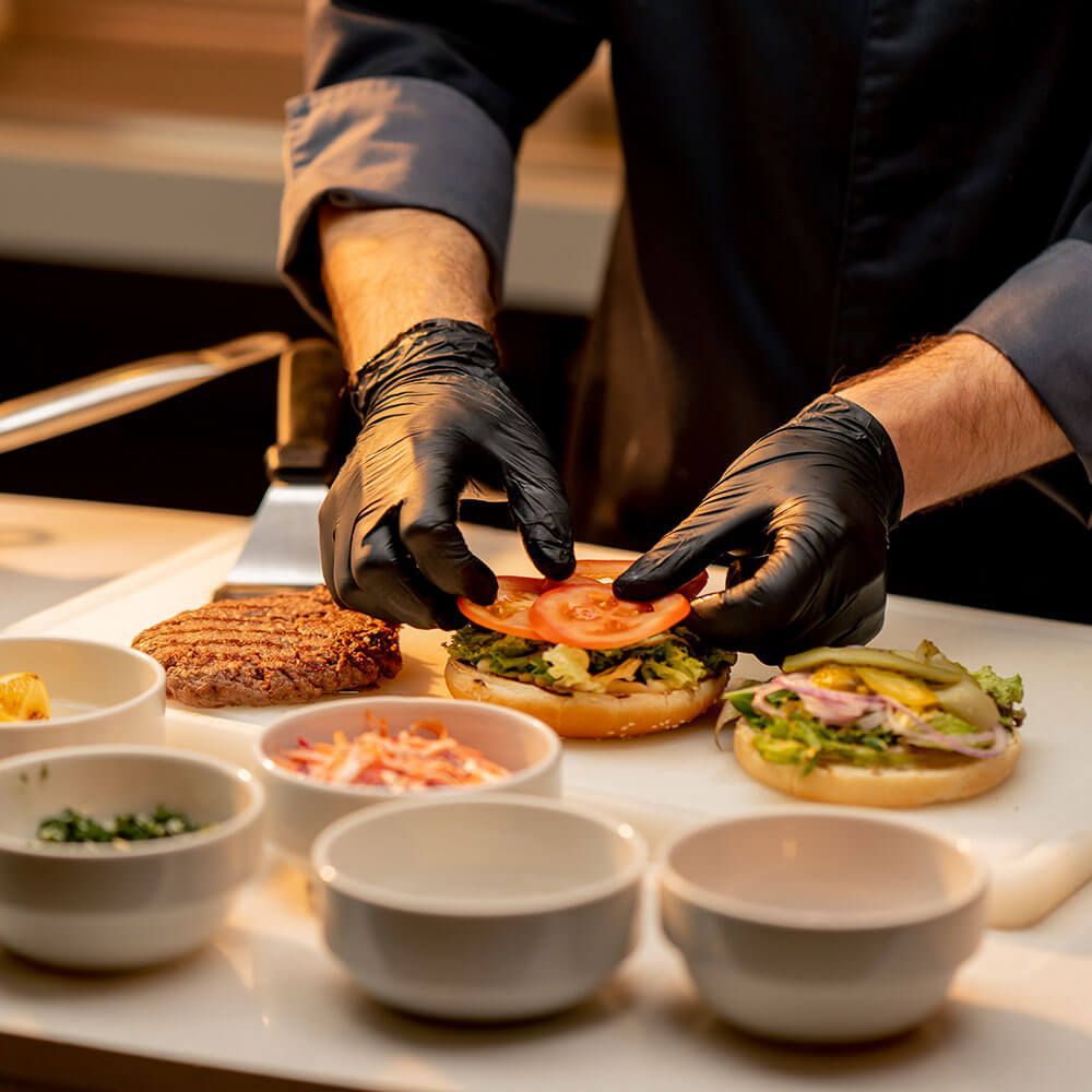 Chef wearing black gloves assembling a burger, with toppings and burger patties visible.