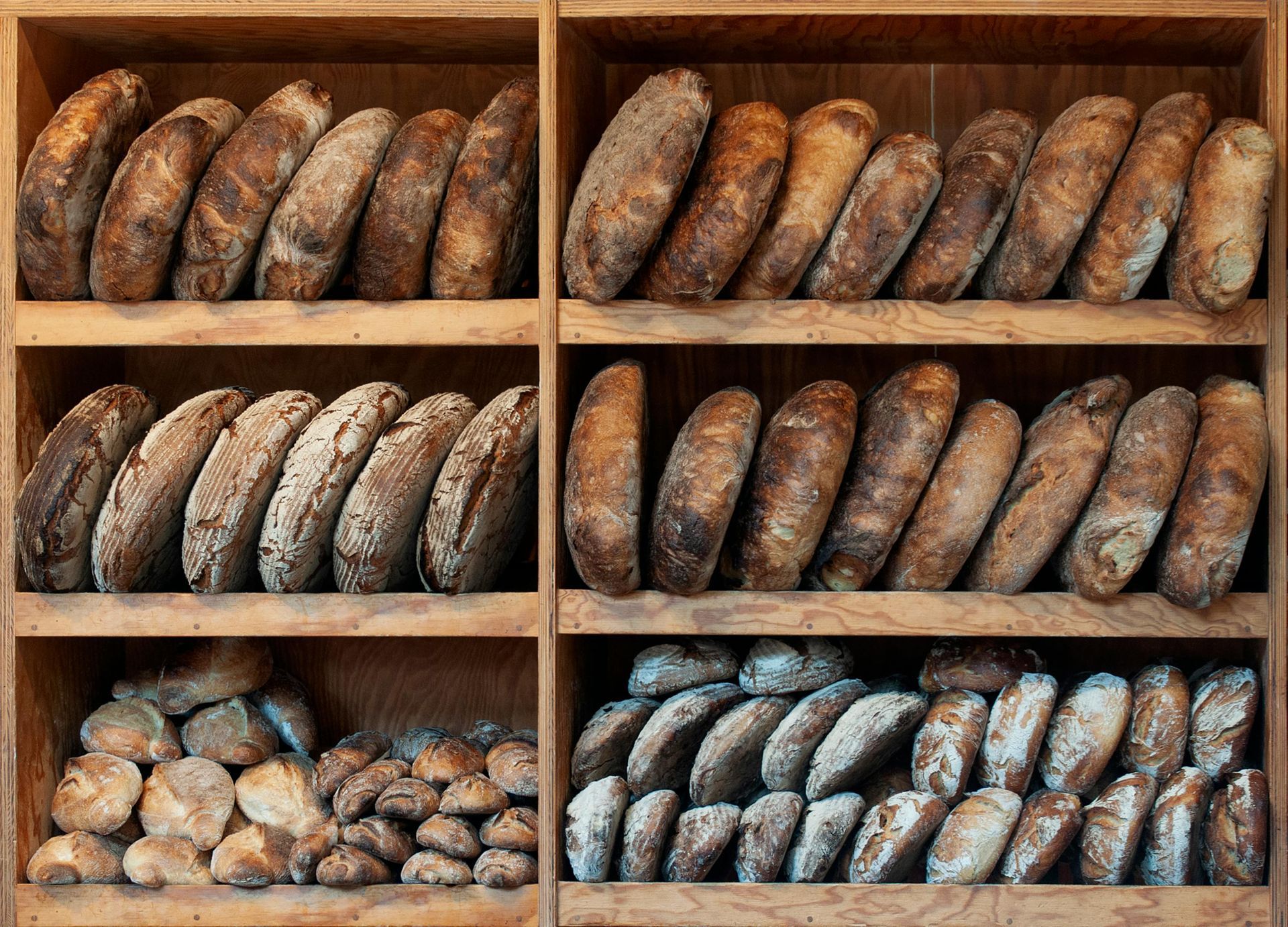 Three wooden shelves filled with rows of round, crusty artisan loaves of bread, arranged in a bakery display.