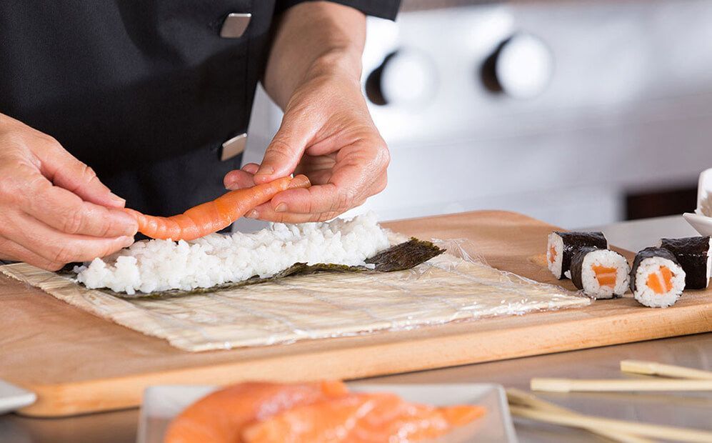 Chef rolling sushi: placing salmon on rice-covered nori. Cut rolls and salmon pieces on the table.