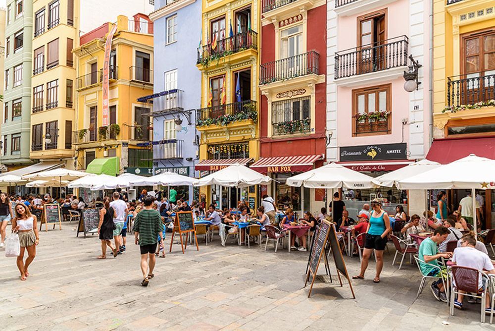 Un grupo de personas están sentadas en mesas bajo sombrillas en una plaza de la ciudad.